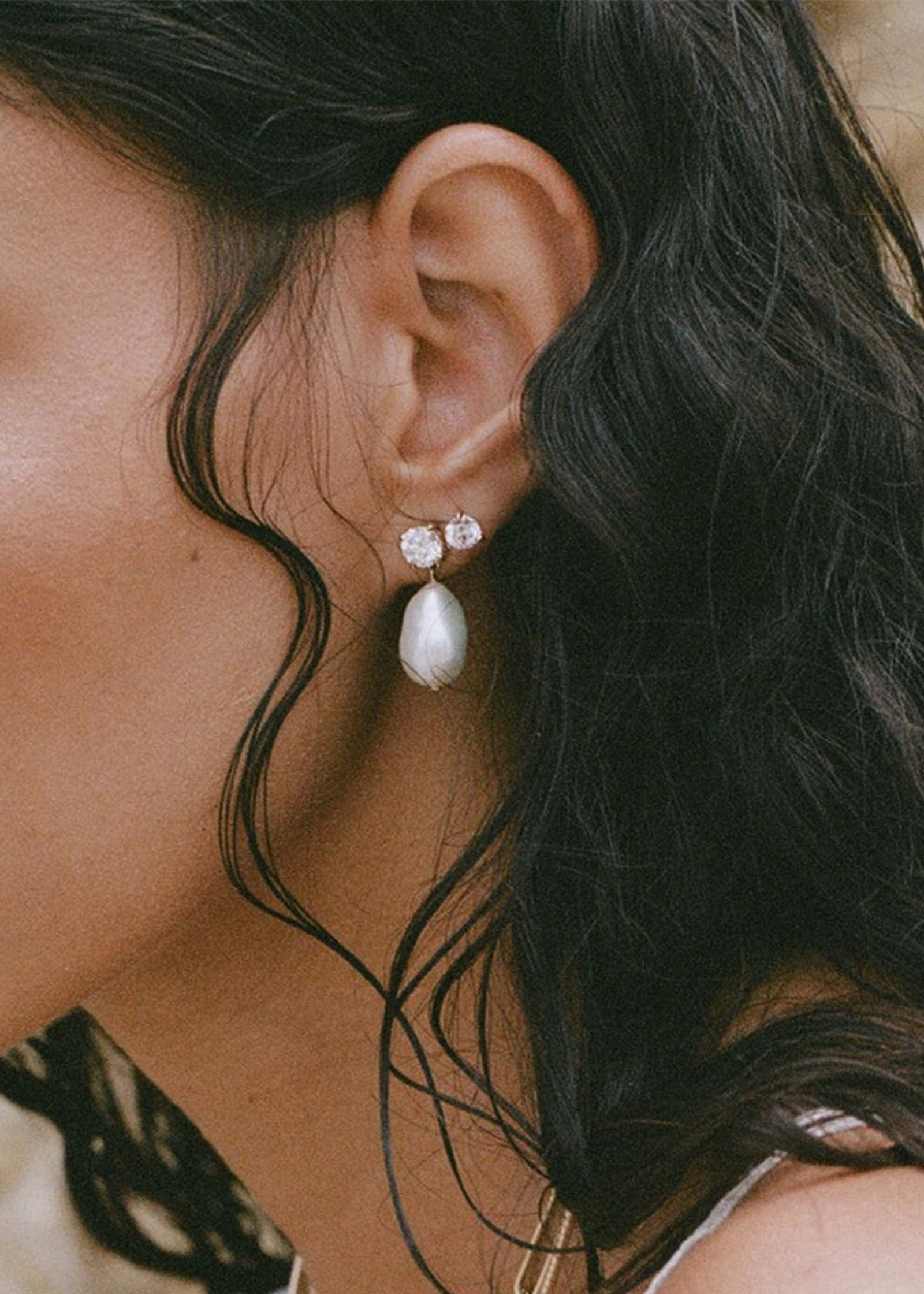 close up of woman's ear wearing Kinn 14k Gold Delanie Diamond Baroque Pearl Earrings layered with June Round Diamond studs in her second piercing