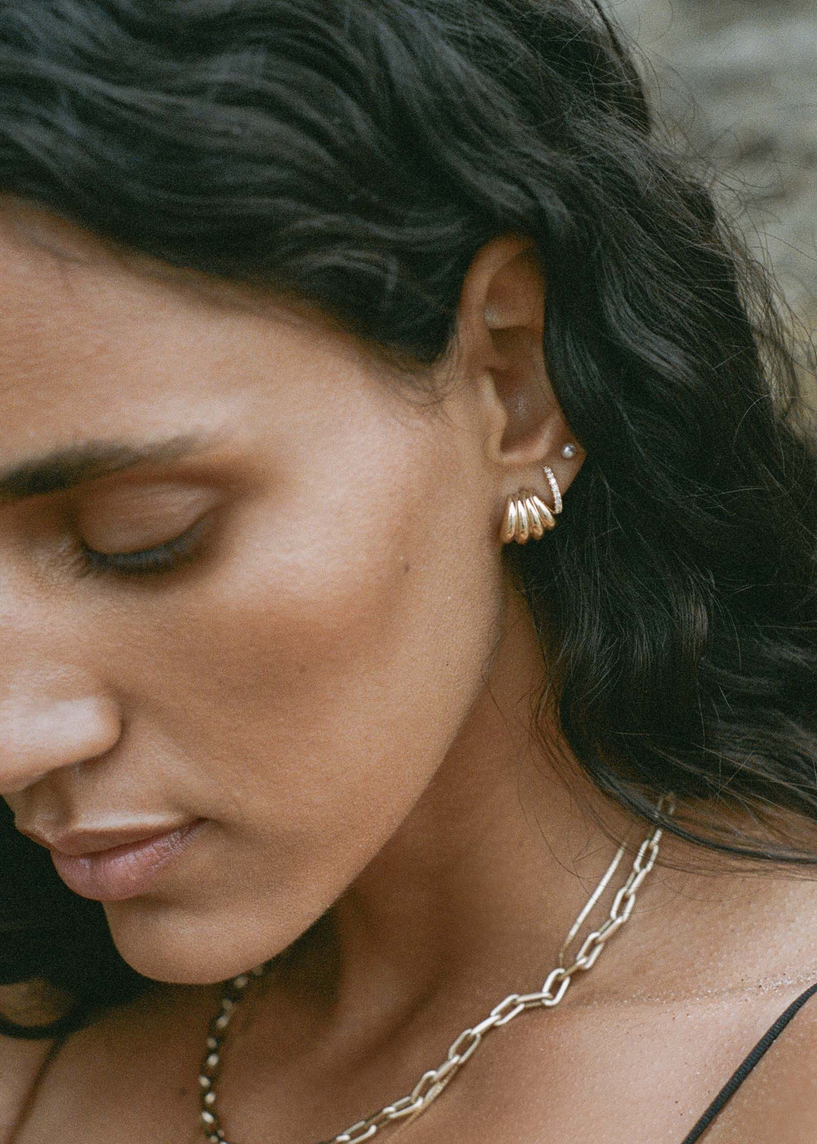close up of a woman's ear wearing Kinn 14k Gold Gaia Scallop Earrings layered with Pave Diamond Huggie Earrings and a Pearl Stud
