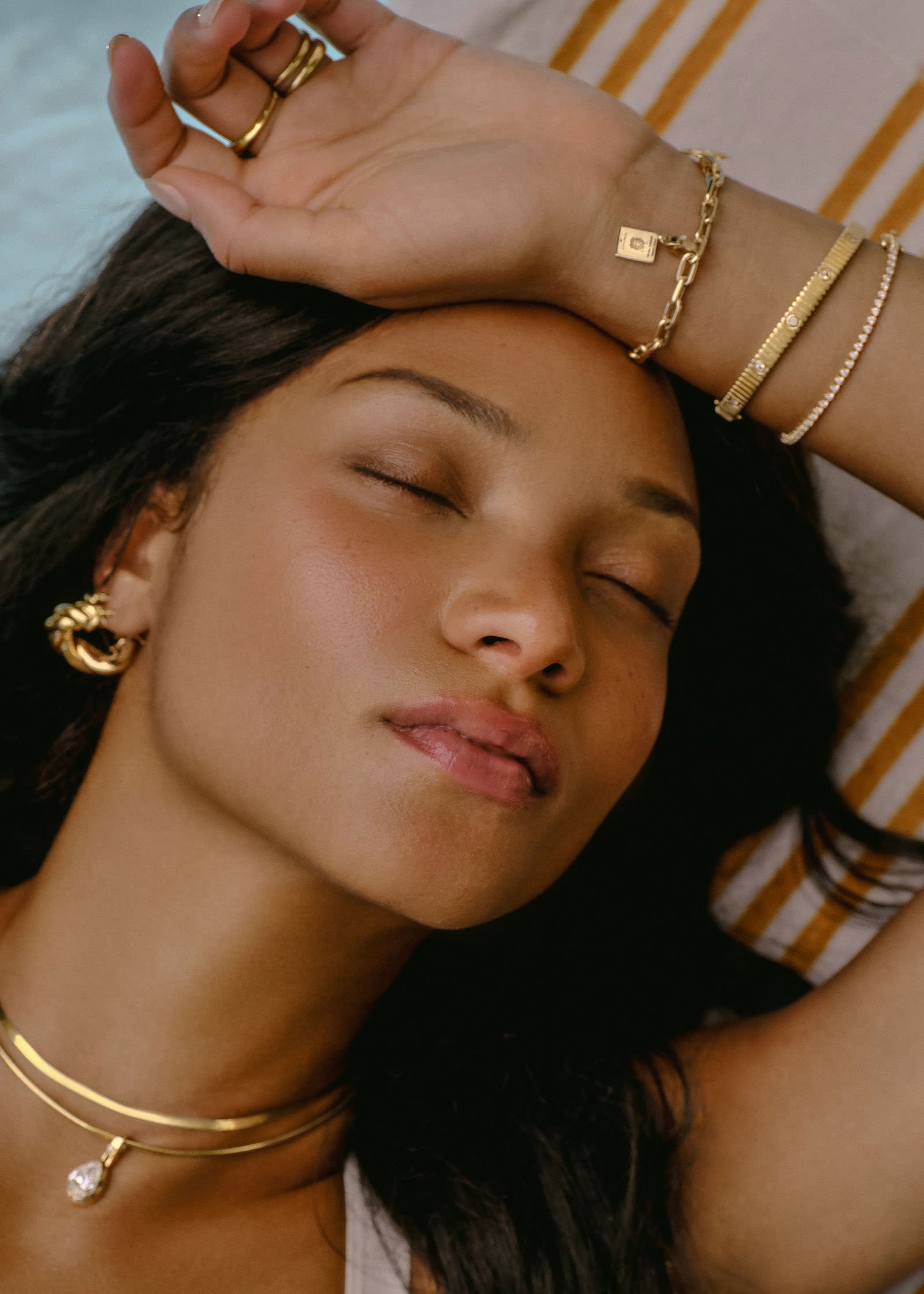 woman laying down with her hand raised to her head wearing a Kinn 14k Gold Italian Heritage Passport Charm on a Mia Link Chain Bracelet stacked with a Solis Ribbed Anniversary Bracelet and Diana Tennis Bracelet