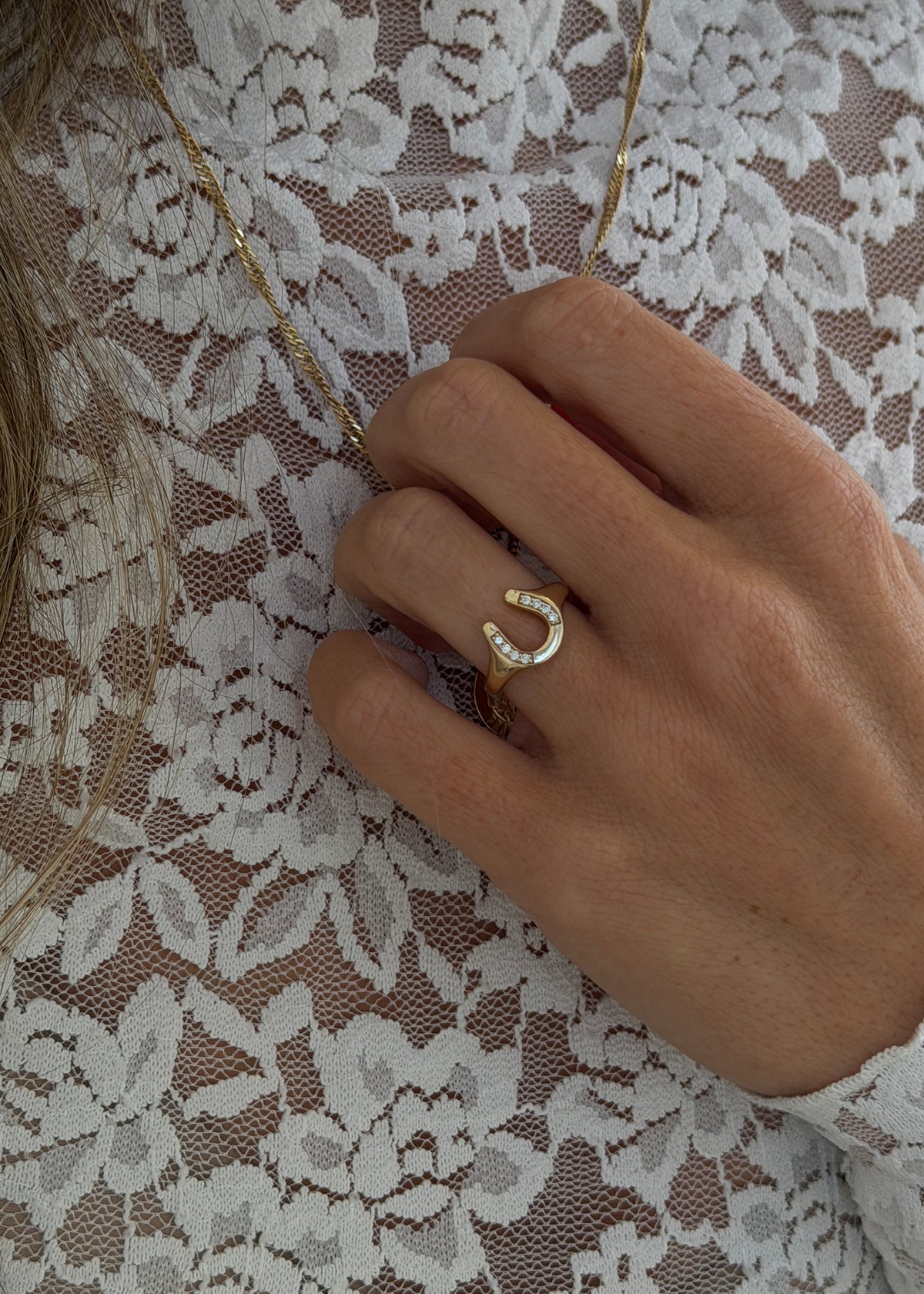 A close-up of a hand wearing a Kinn 14k gold horseshoe ring with small clear stones, layered over a white lace top.