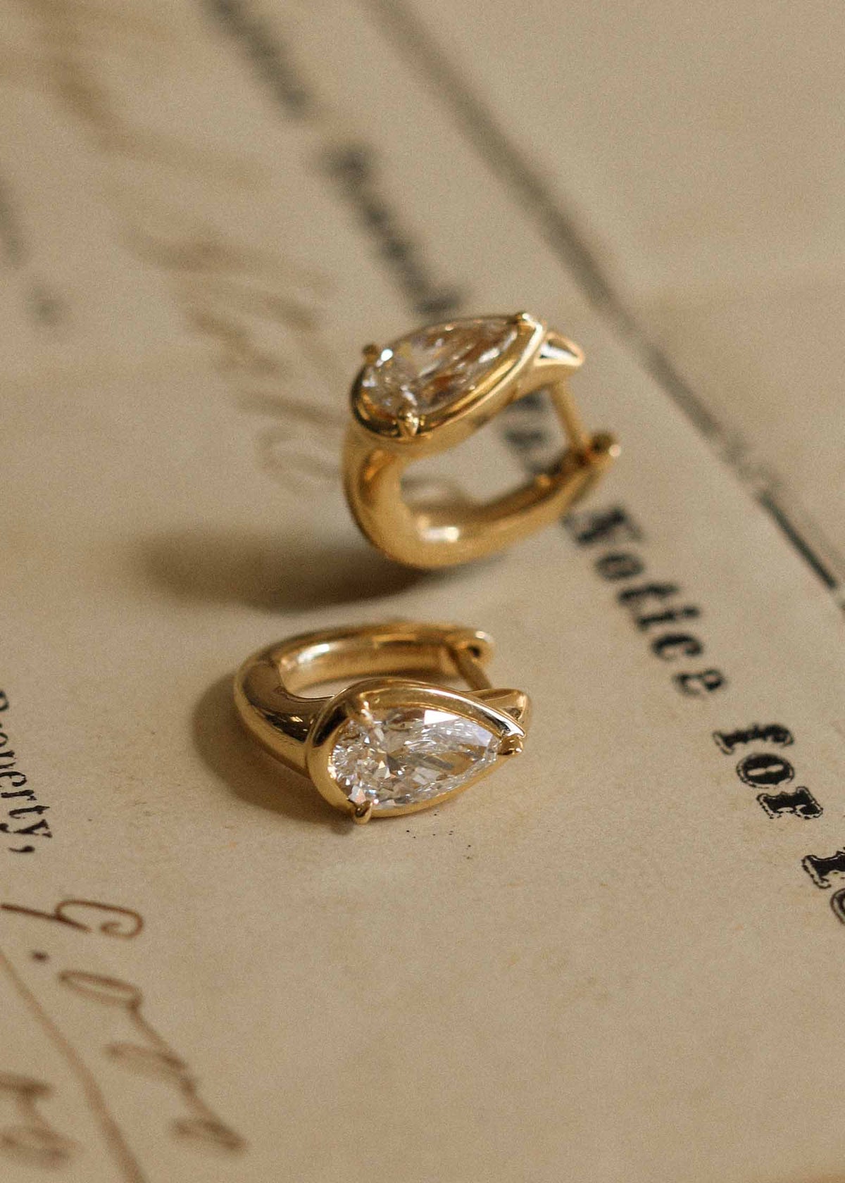 close up of a pair of Kinn 14k Gold Jackson Pear Diamond Huggie Hoop Earrings laying against old letters