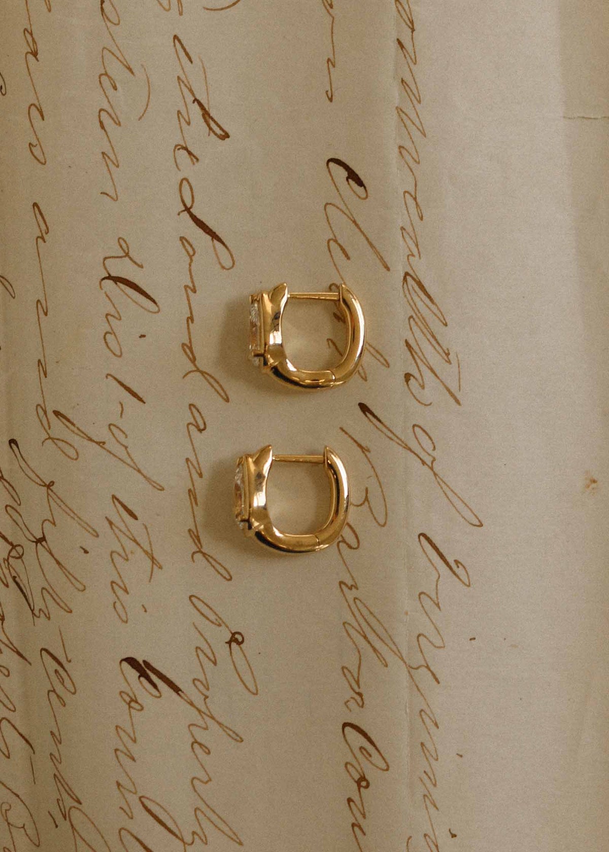 A pair of Kinn 14k Gold Jackson Pear Diamond Huggie Hoop Earrings laying on their side flat against an old handwritten letter