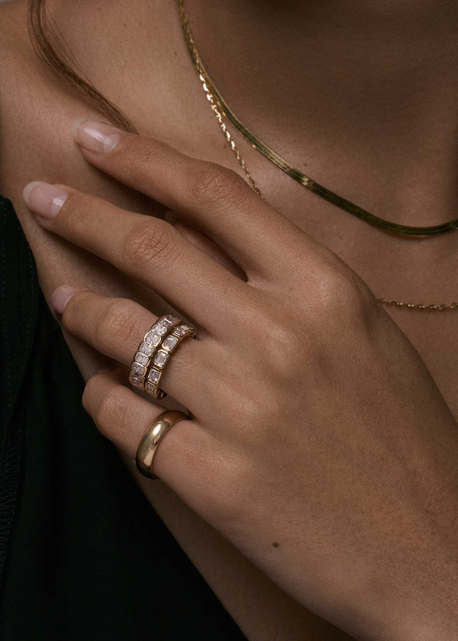 close up of a woman's hand wearing a Kinn Studio 14k Gold Jane Radiant Diamond Eternity Band Ring stacked with a Marianna Emerald Cut Diamond Bezel Eternity Band Ring on her ring finger and a Milo Wedding Band on her pinky