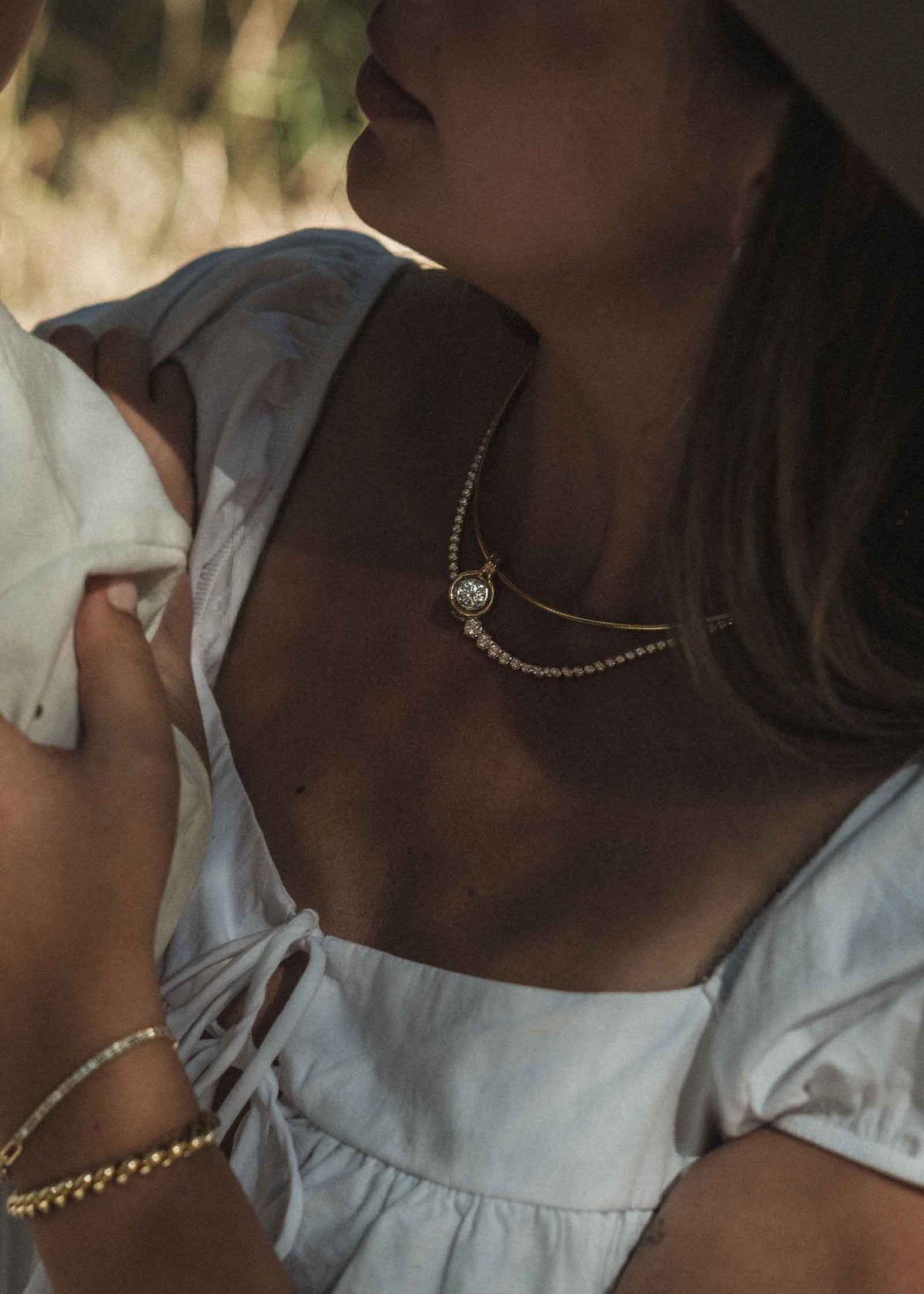 woman in a white dress wearing a Kinn Studio 14k Gold Jody Round Diamond Pendant on an Heirloom Curved Chain Necklace layered with a Naomi Diamond Tennis Necklace