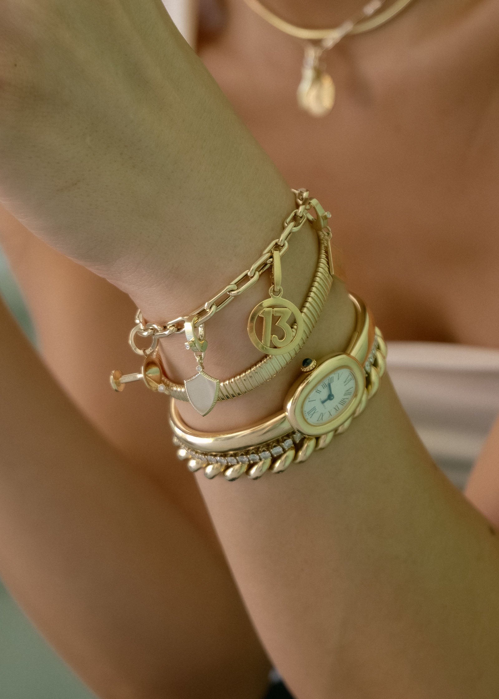 A close up of a woman's arm wearing a Kinn 14k Gold Dirty Martini Charm Diamond, Ace Shield Charm, and Vintage Cartier 13 Charm hanging off a Mia Link Chain Bracelet and stacked with other gold and diamond bracelets and a vintage watch