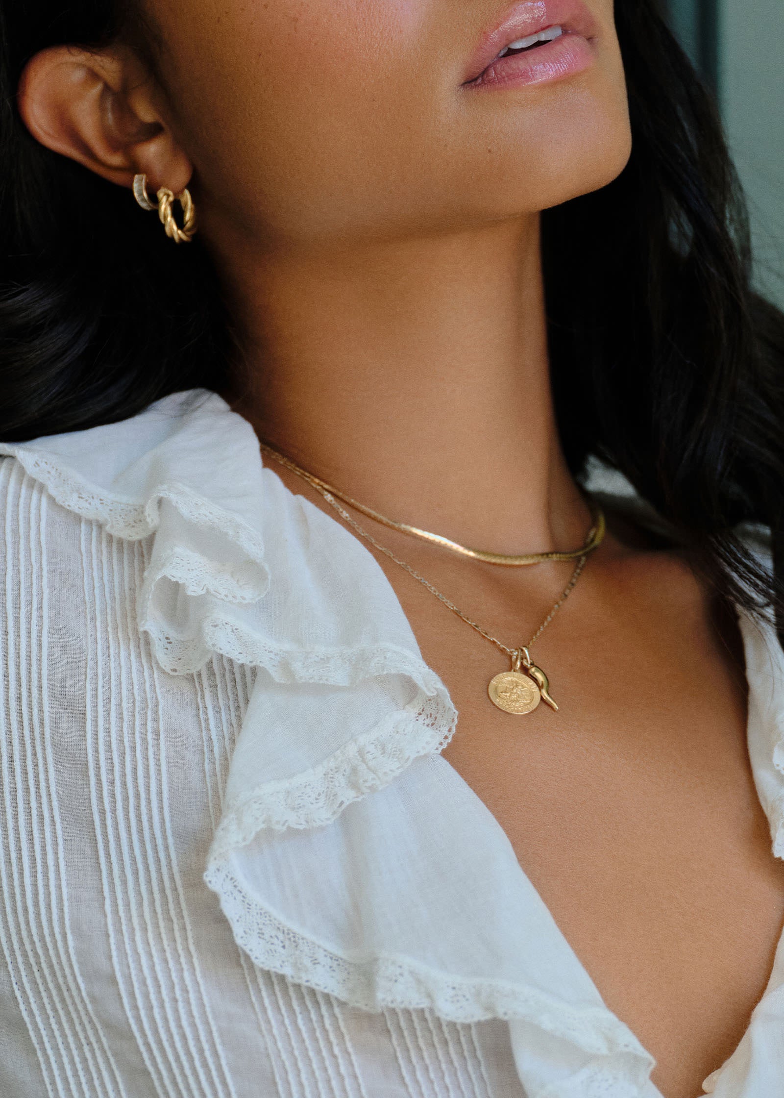 A woman in a white ruffled shirt wearing a Kinn 14k Gold Saint Michael Charm Pendant and Alessandra Cornicello Charm hanging off a Valentino Tri Color Gold Chain layered with a Petite Kennedy Herringbone chain and wearing Petite Taylor Twist Hoops