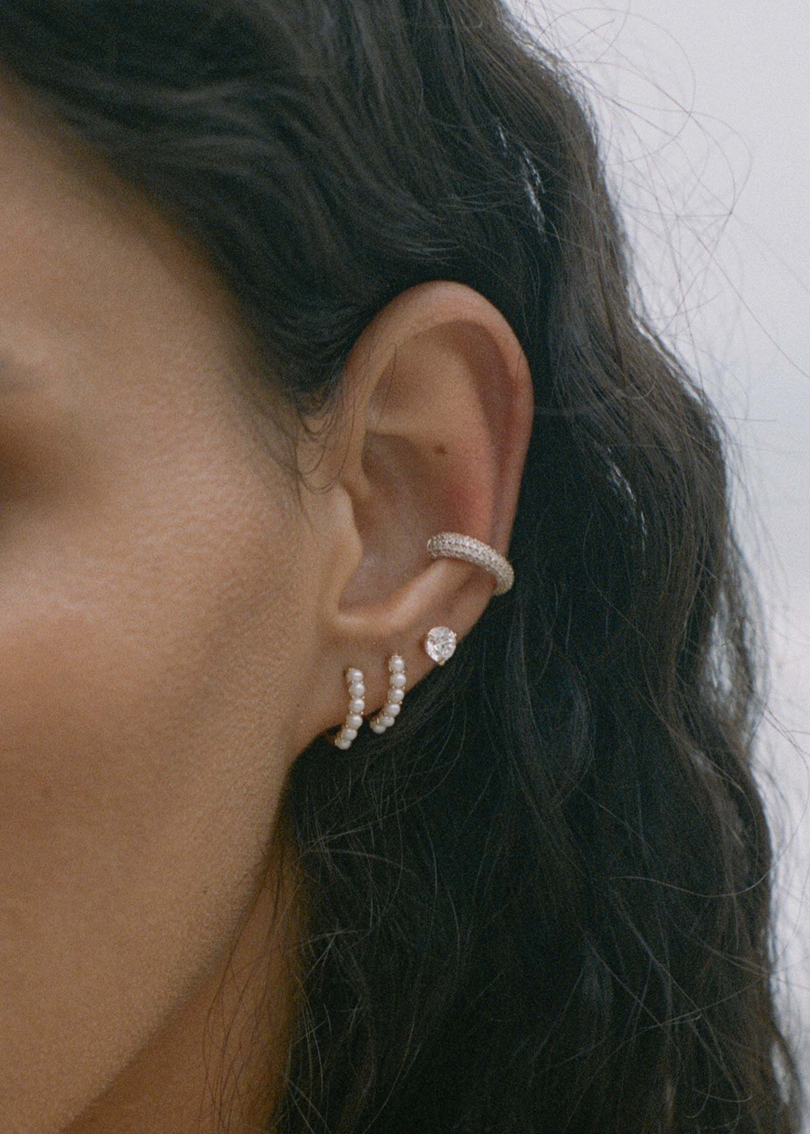 close up of a woman's ear wearing two Kinn 14k Gold Marina Pearl Huggie Hoop Earrings layered together in her first and second piercing along with a June Round Stud and a Love Pave Diamond ear cuff