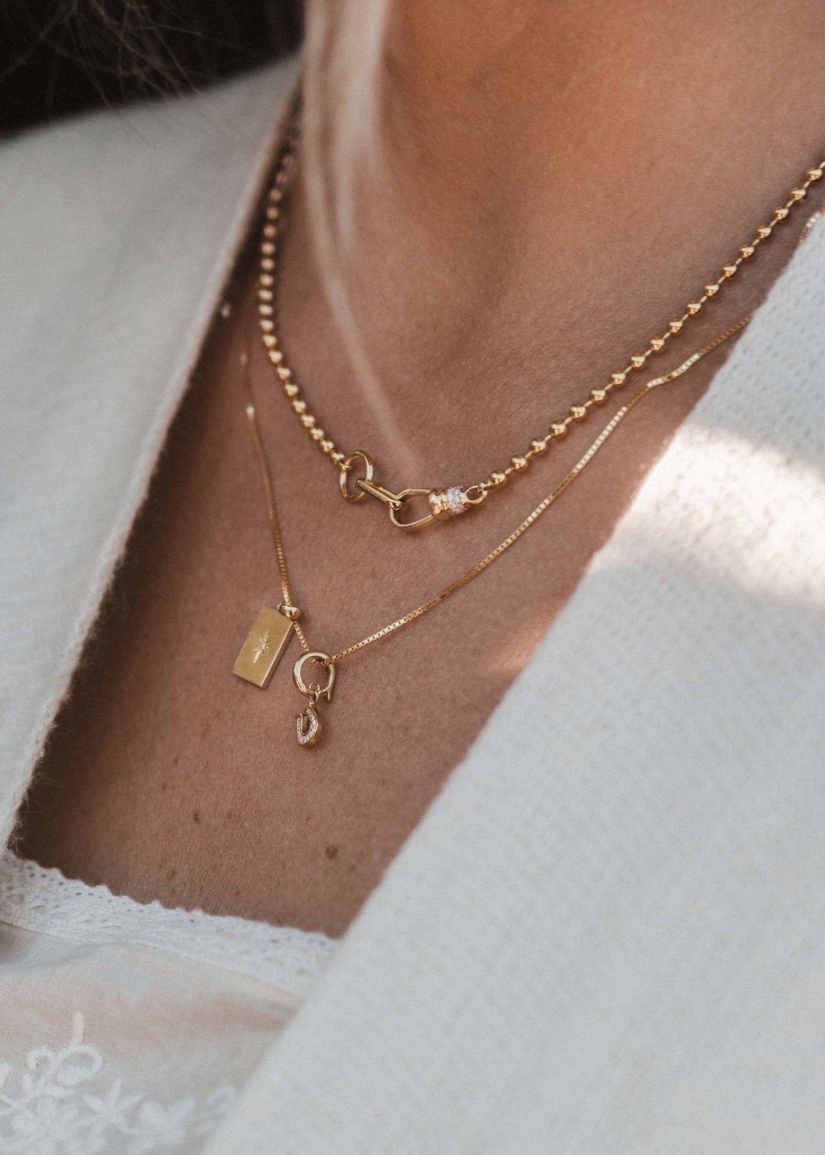 A close up of a woman wearing a Kinn 14k Gold Mini Horseshoe Baguette Diamond Charm and North Star Pendant Diamond on a box chain necklace layered with a Pave Diamond Ball Chain Necklace