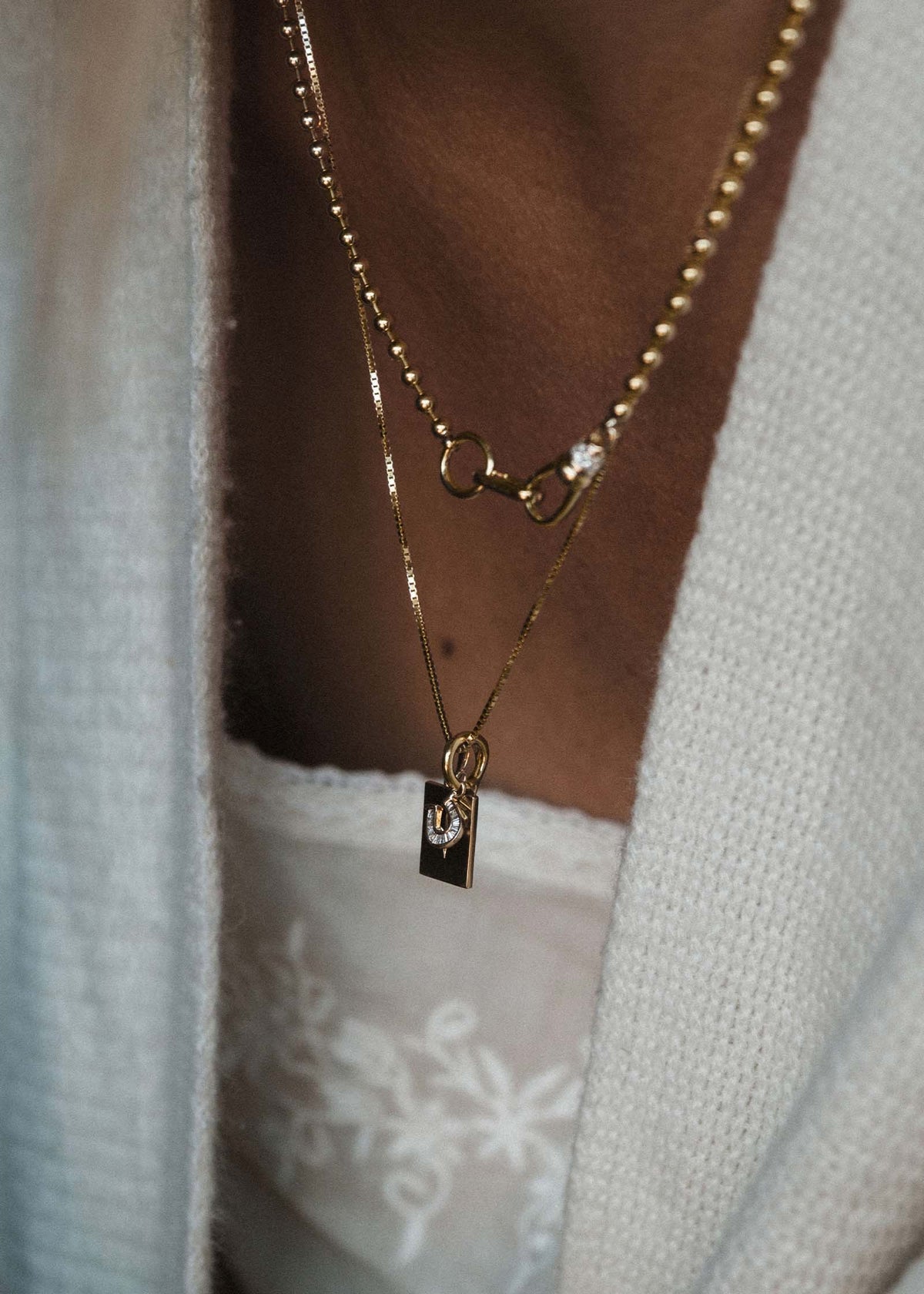 A close up of a woman wearing a Kinn 14k Gold Mini Horseshoe Baguette Diamond Charm and North Star Pendant Diamond on a box chain necklace layered with a Pave Diamond Ball Chain Necklace