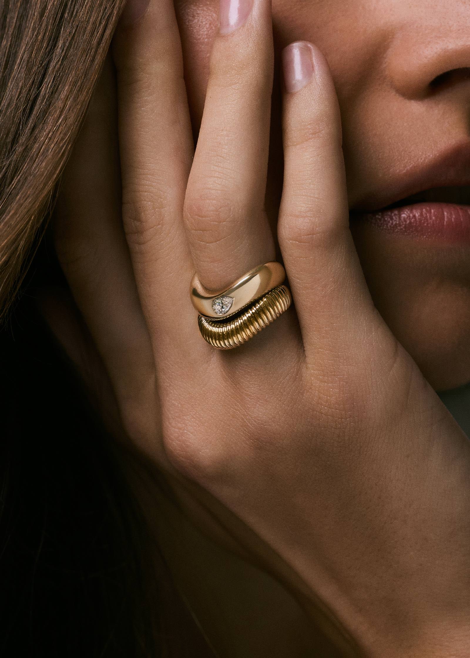 A Kinn 14k Gold Demi Curve Band Ring with a Pear Diamond and a Demi Coil Ribbed Band Ring stacked together on a woman's ring finger with her hand resting against her face