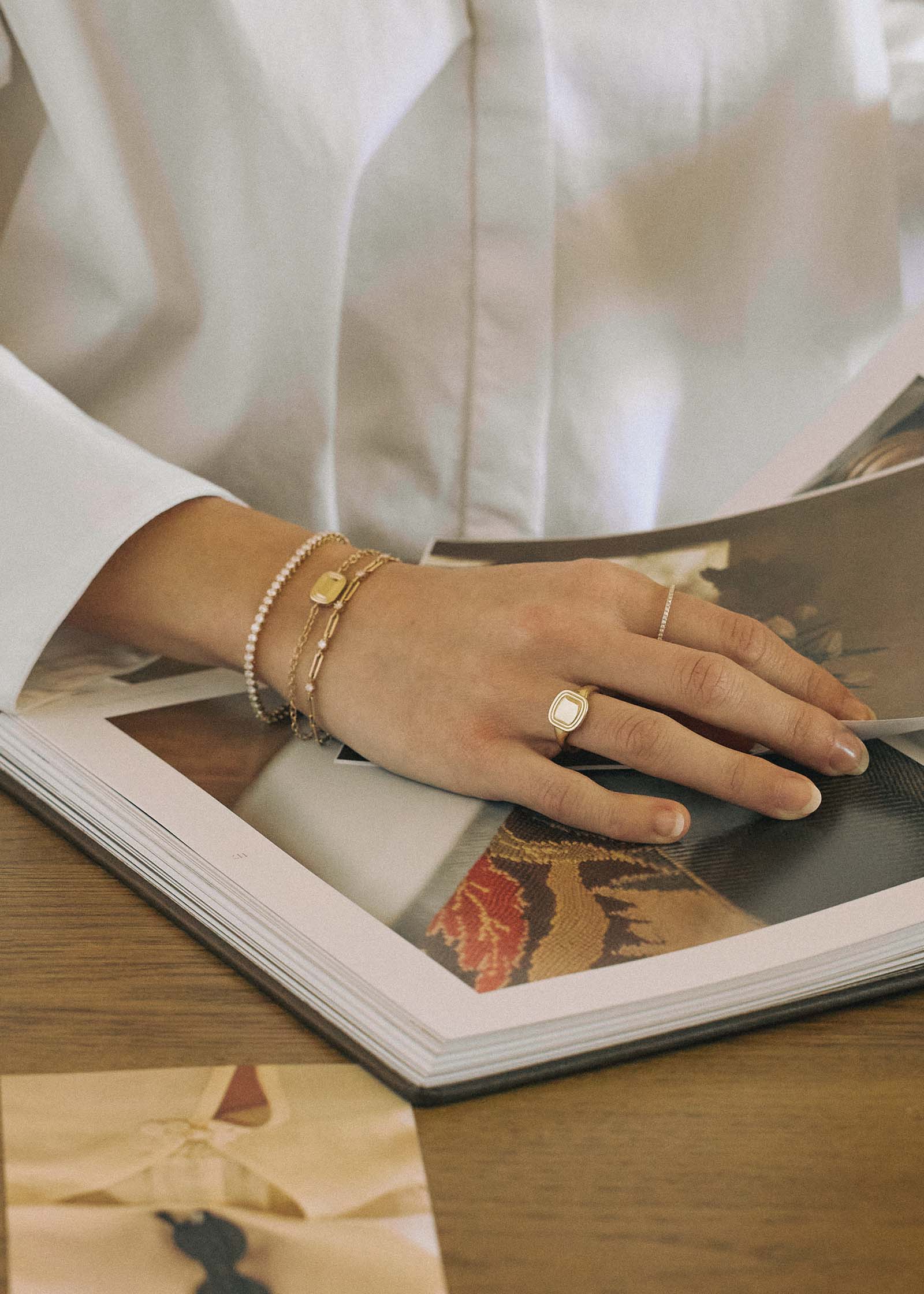 woman with her hand laying flat on an open book wearing a Kinn x Shoppe Amber Interiors The Estate Bracelet 14k Yellow Gold stacked with a North Star Anniversary Diamond Bracelet and a Diamond Tennis Bracelet