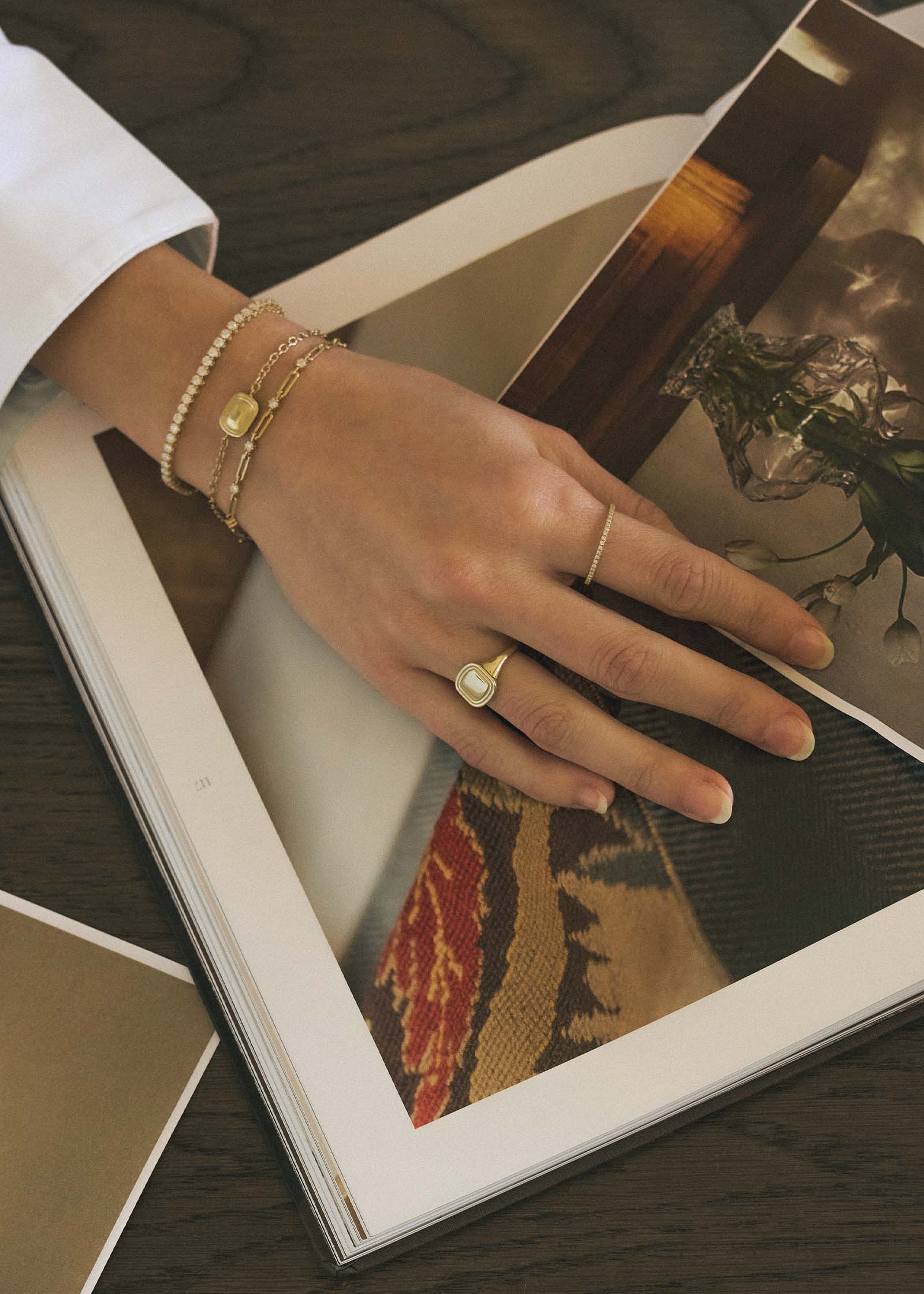 woman with her hand laying flat on an open book wearing a Kinn x Shoppe Amber Interiors The Estate Bracelet 14k Yellow Gold stacked with a North Star Anniversary Diamond Bracelet and a Diamond Tennis Bracelet