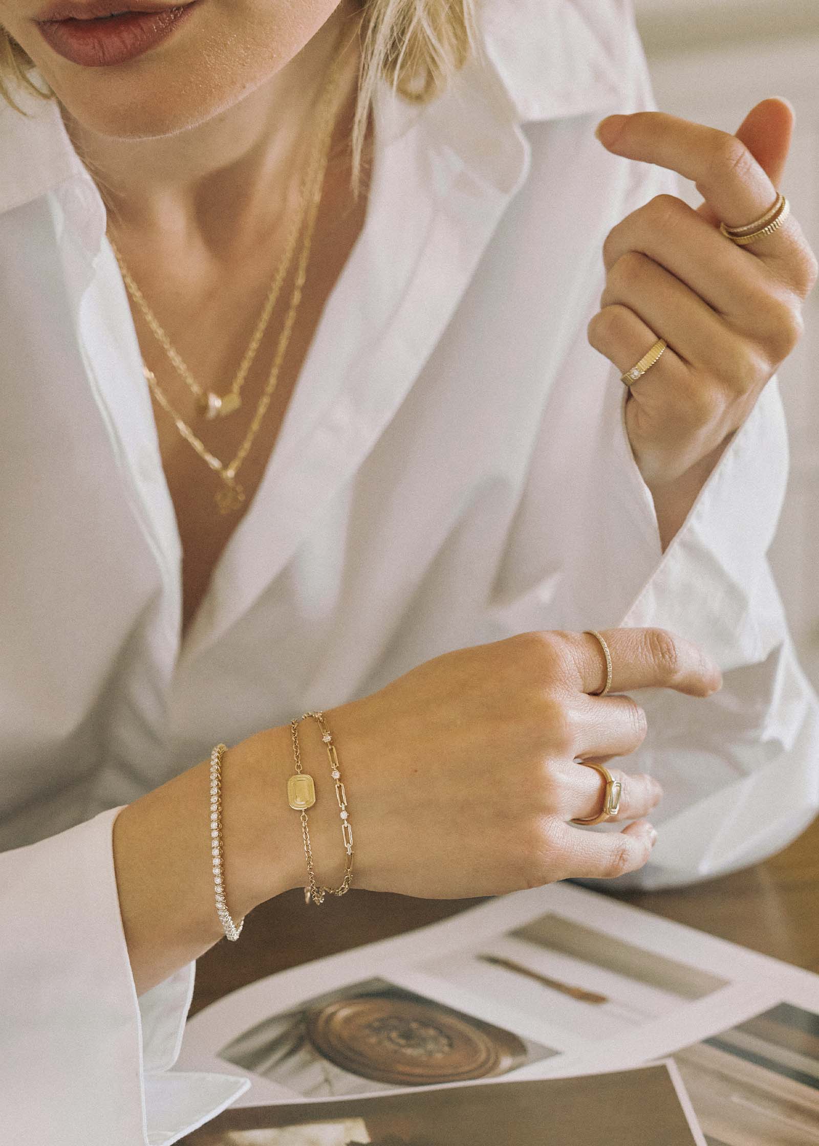 A woman in a white shirt sitting at a desk wearing a Kinn x Shoppe Amber Interiors The Estate Bracelet 14k Yellow Gold stacked with a North Star Anniversary Diamond Bracelet and a Diamond Tennis Bracelet