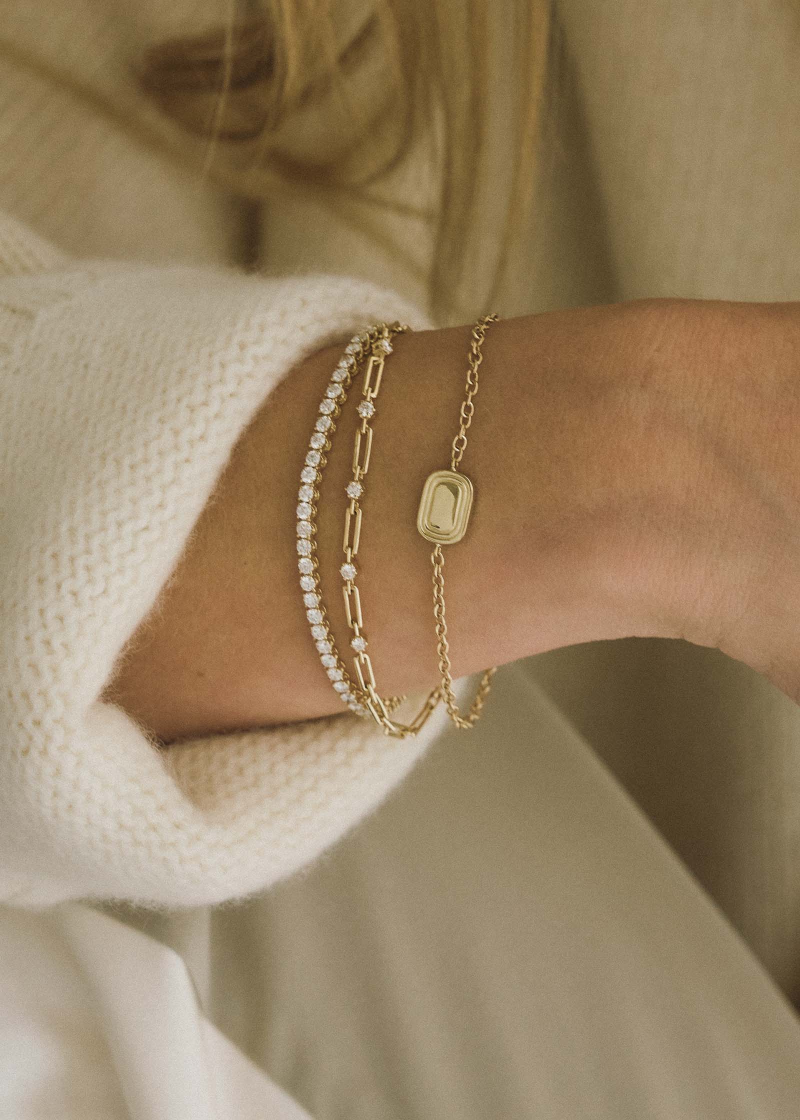 A close up of a woman's wrist wearing a Kinn x Shoppe Amber Interiors The Estate Bracelet 14k Yellow Gold stacked with a North Star Anniversary Diamond Bracelet and a Diamond Tennis Bracelet