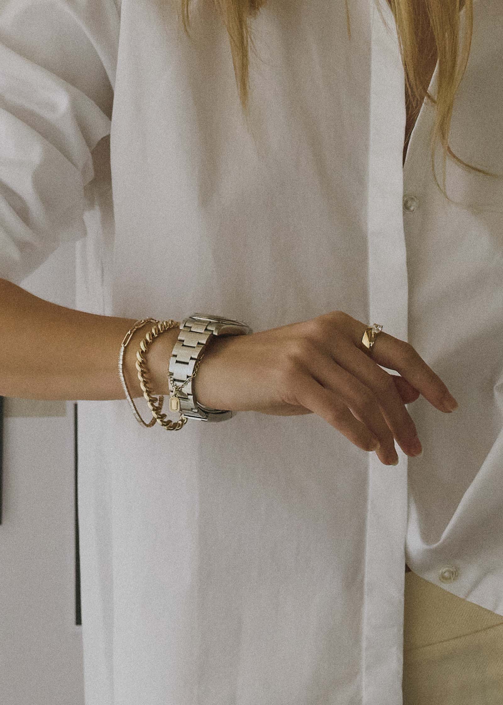 woman's wrist wearing a watch with a Kinn x Shoppe Amber Interiors The Estate Watch Charm 14k Yellow Gold dangling from the band and stacked with a Hampshire House 14k Gold Bracelet and a Serena Baguette Diamond Bracelet