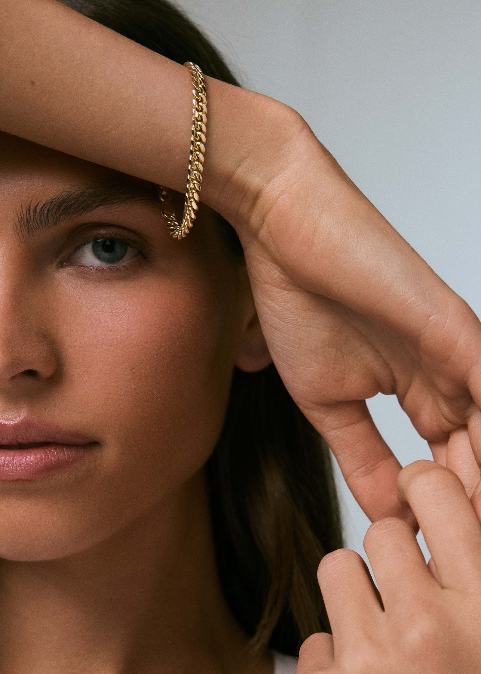 A woman with her arm draped over her head wearing a Kinn 14k Gold Familia Cuban Chain Bracelet
