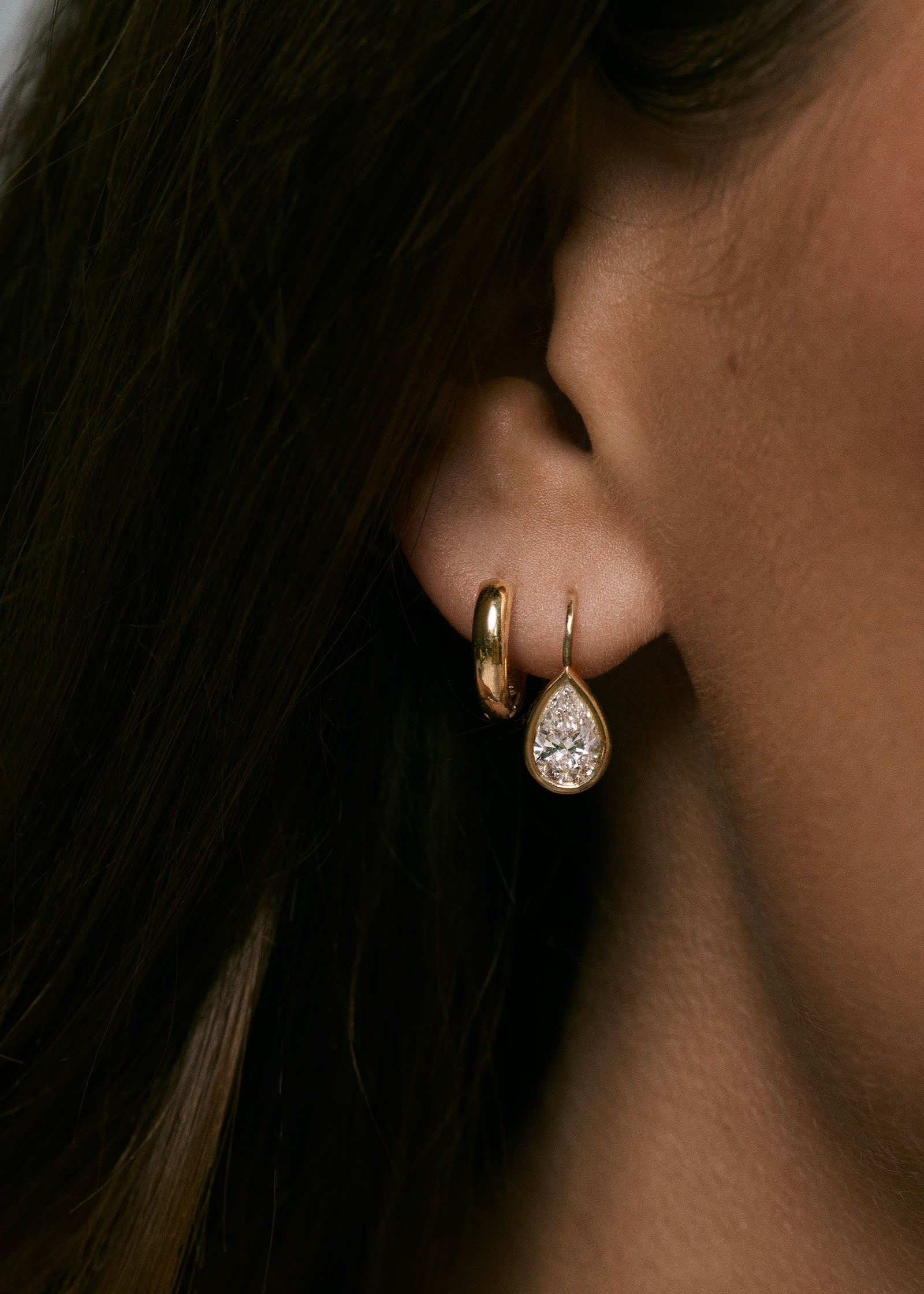 A brunette woman wearing a pair of Kinn 14k Gold 2ctw Pear Diamond Drop Earrings layered with a huggie hoop