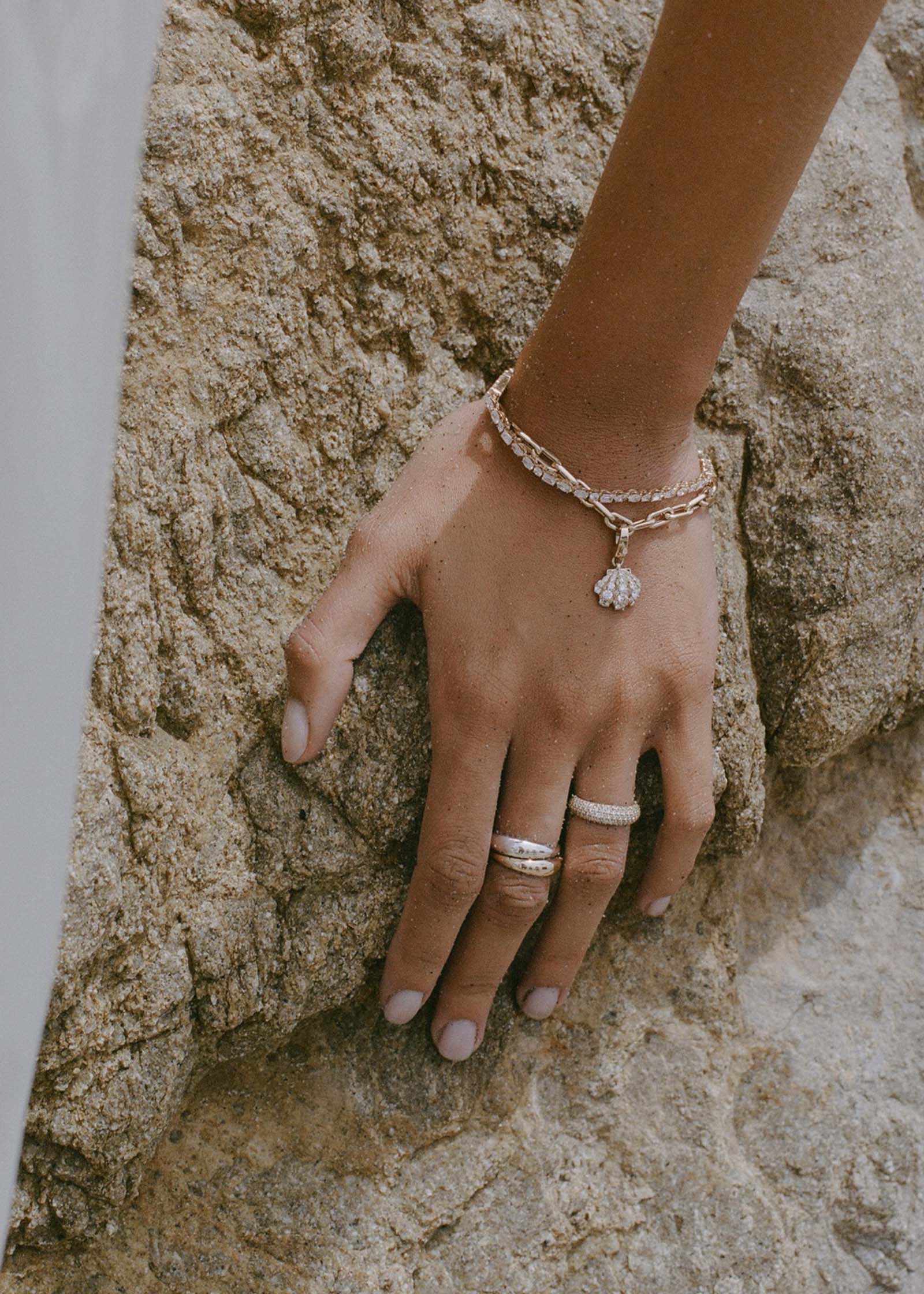 woman's hand resting against a rock wearing a Kinn 14k Gold Pavé Diamond Shell Charm Bracelet and an Esme Diamond Tennis bracelet stacked on her wrist and two Dare to Love Dome Rings and a Scarlett Pave Diamond Ring stacked on her fingers