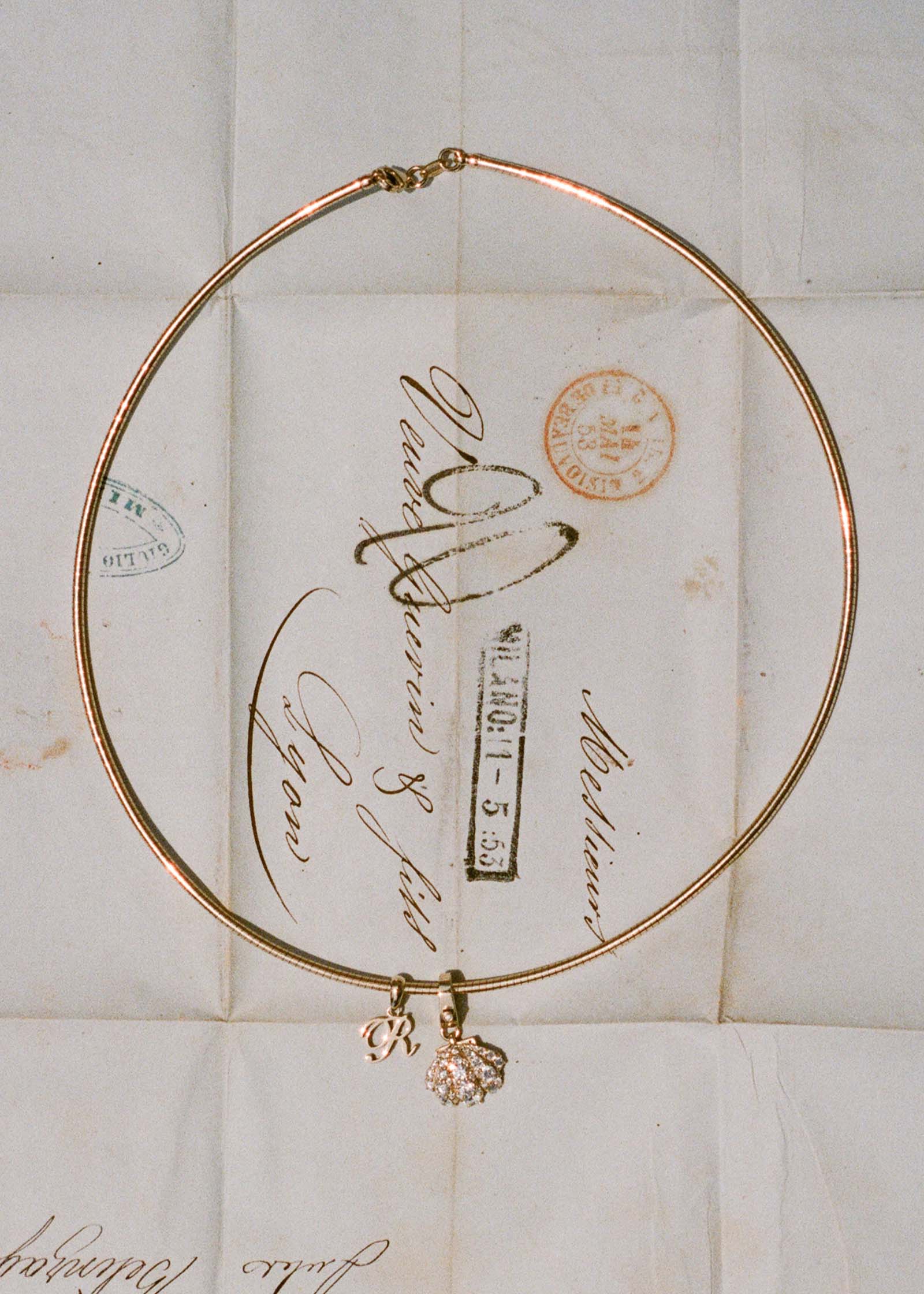 A Kinn 14k Gold Pavé Diamond Shell Charm Pendant and a Love Letter R Charm on an Heirloom Curved Chain Necklace against a unfolded stamped letter
