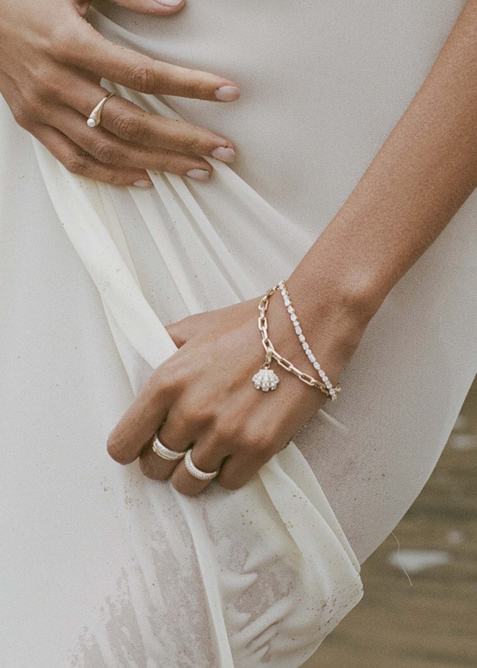 woman in a white dress on the beach wearing a Kinn 14k Gold Pavé Diamond Shell Charm Bracelet and an Esme Diamond Tennis bracelet stacked on her wrist and a Pearl Signet Ring on her other hand