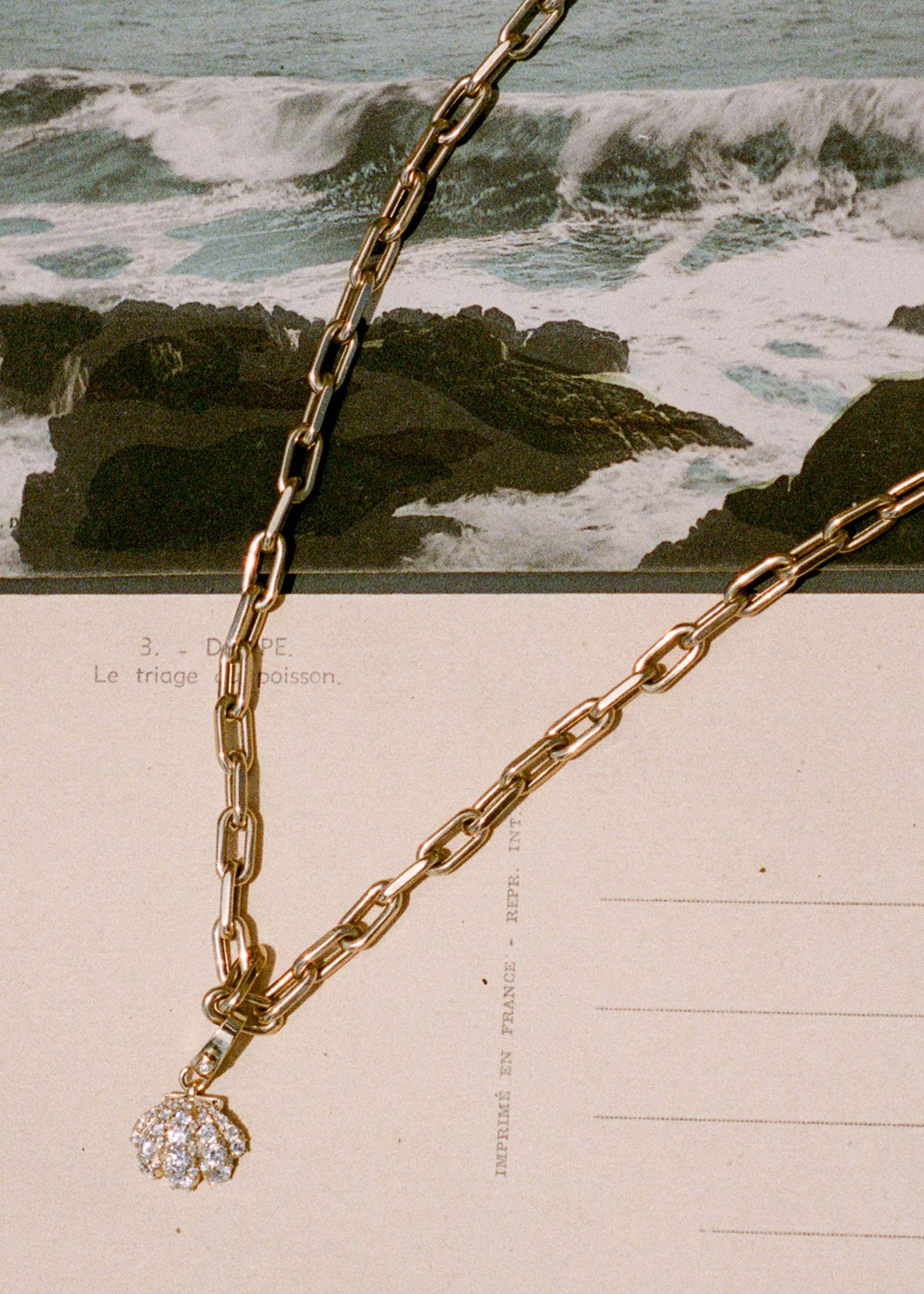a Kinn 14k Gold Pavé Diamond Shell Charm Necklace on Mia Link Chain laying flat against a postcard and picture of the ocean
