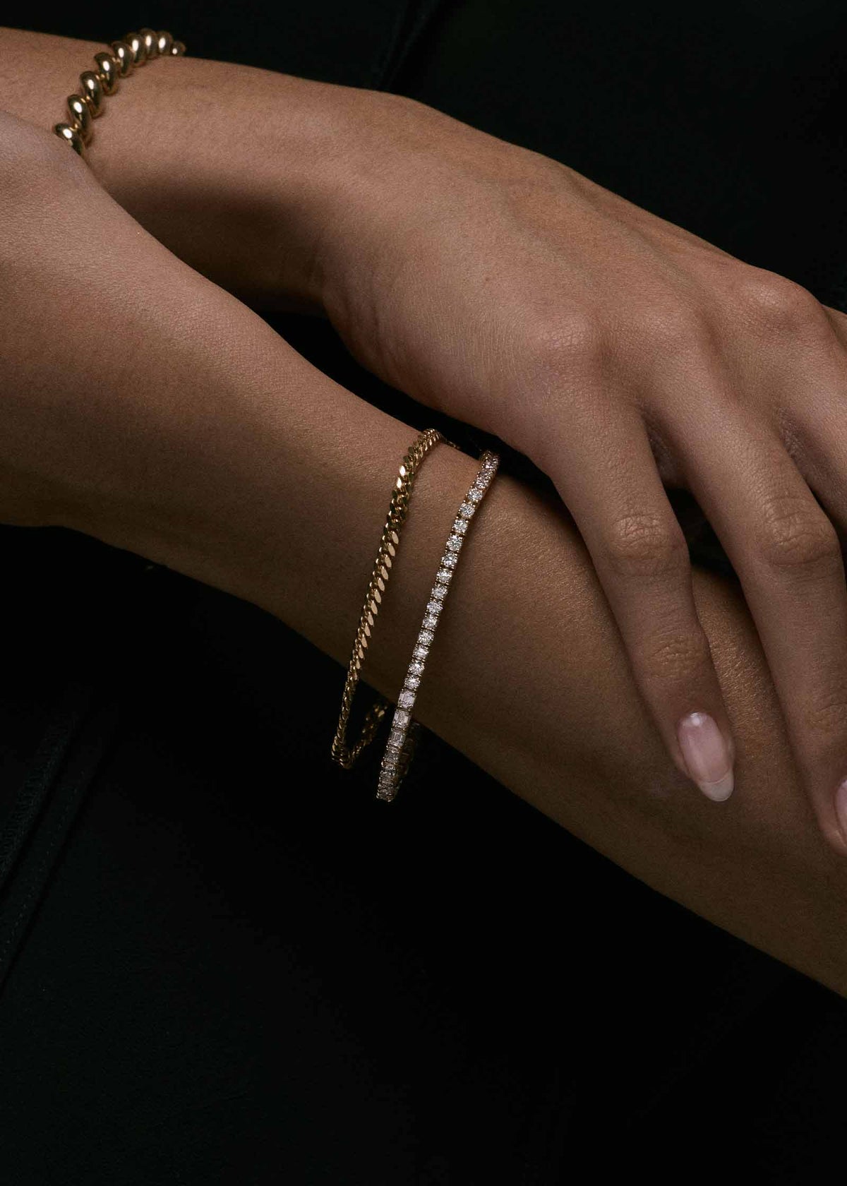 A close-up of a person's wrist and hand wearing a Kinn x Prince 14k Gold Billie Half Round Emerald Tennis Bracelet stacked with a Petite Capri Curb Chain Bracelet
