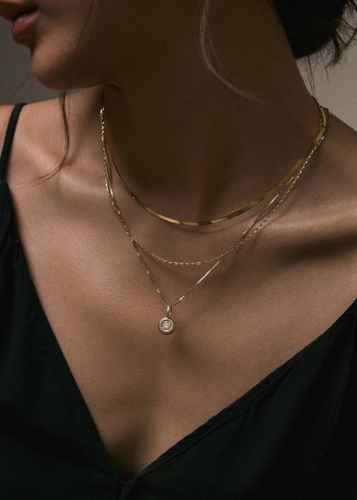 A woman in a black top wearing a Kinn 14k Gold Micro Mariner Chain Necklace layered with a Petite Carter Herringbone Chain and a Delphine Ribbed Diamond Pendant necklace