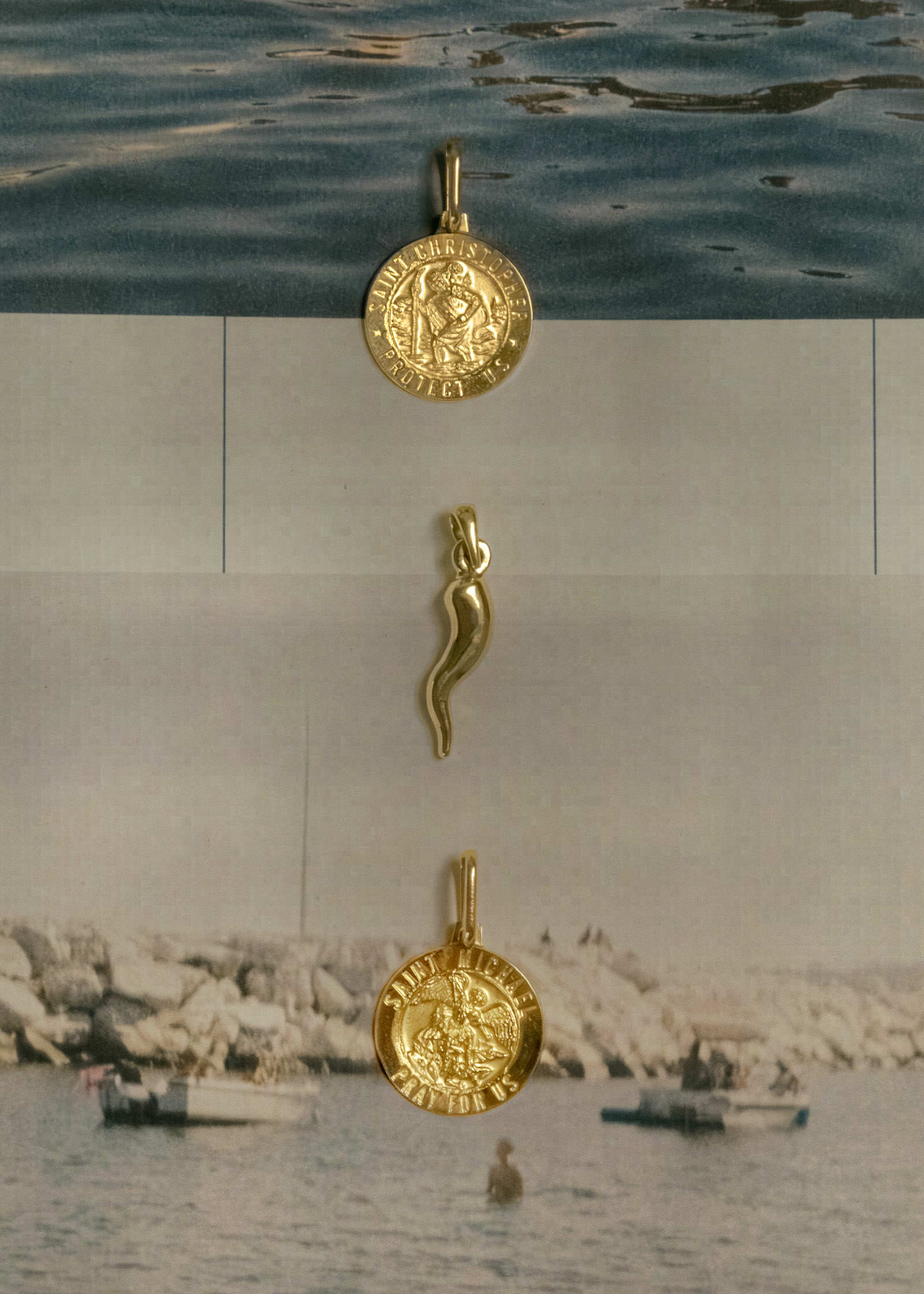 A Kinn 14k Gold Saint Christopher Charm Pendant, Alessandra Cornicello Charm, and a Saint Michael Charm Pendant laying flat in a vertical line on top of photos of Italy and the Italian coast