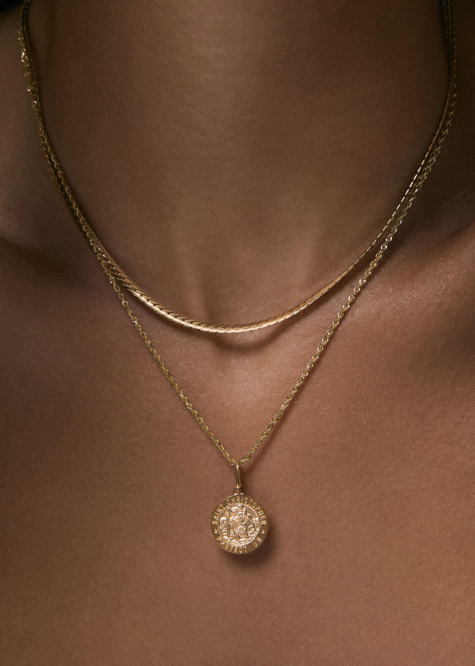 A close up of a woman wearing a Kinn 14k Gold Saint Christopher Charm Pendant on a Petite Rope Chain Necklace layered with a Petite Kennedy Herringbone Chain Necklace