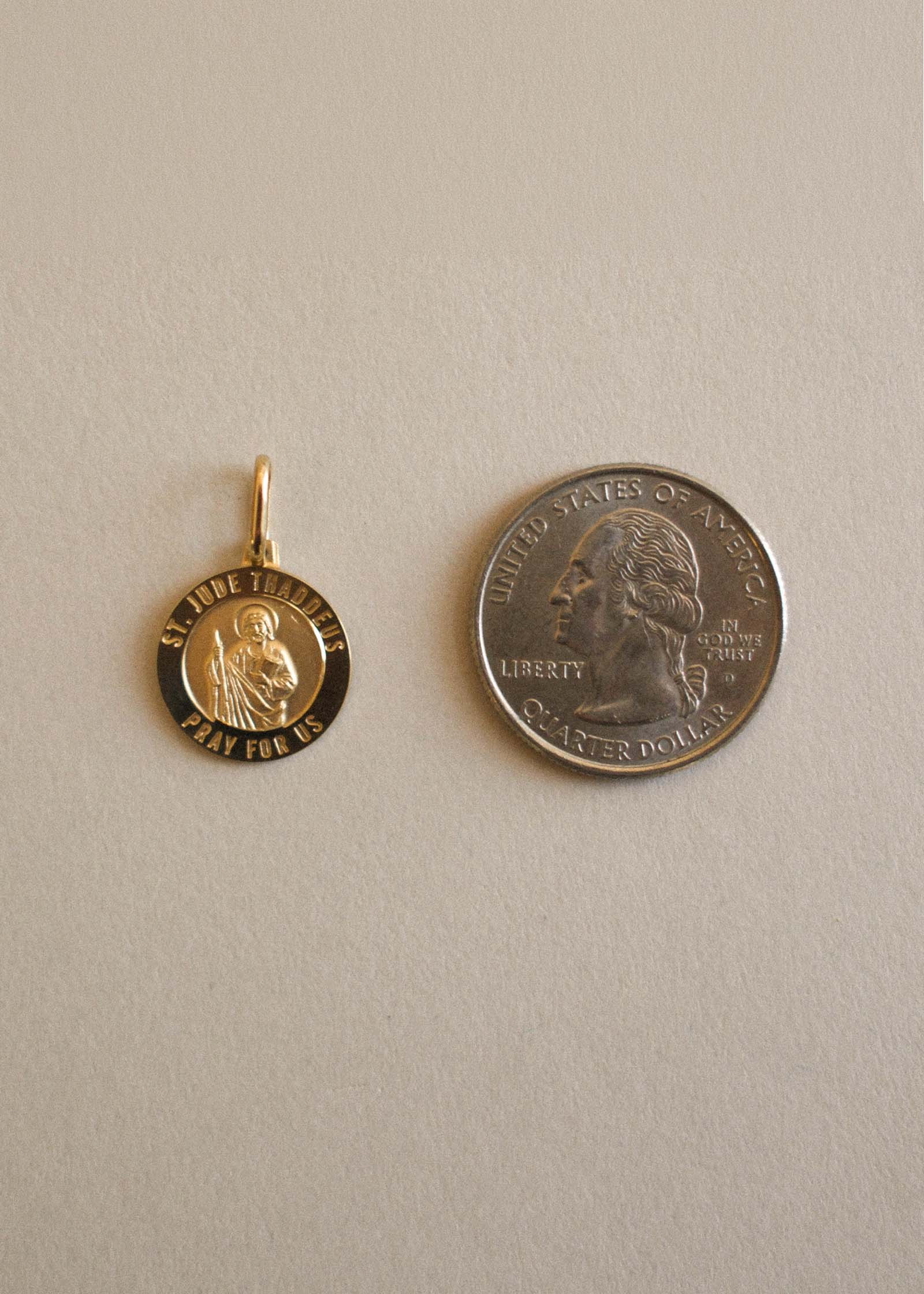 A size comparison flat lay of a Kinn 14k Gold Saint Jude Charm Pendant next to a quarter