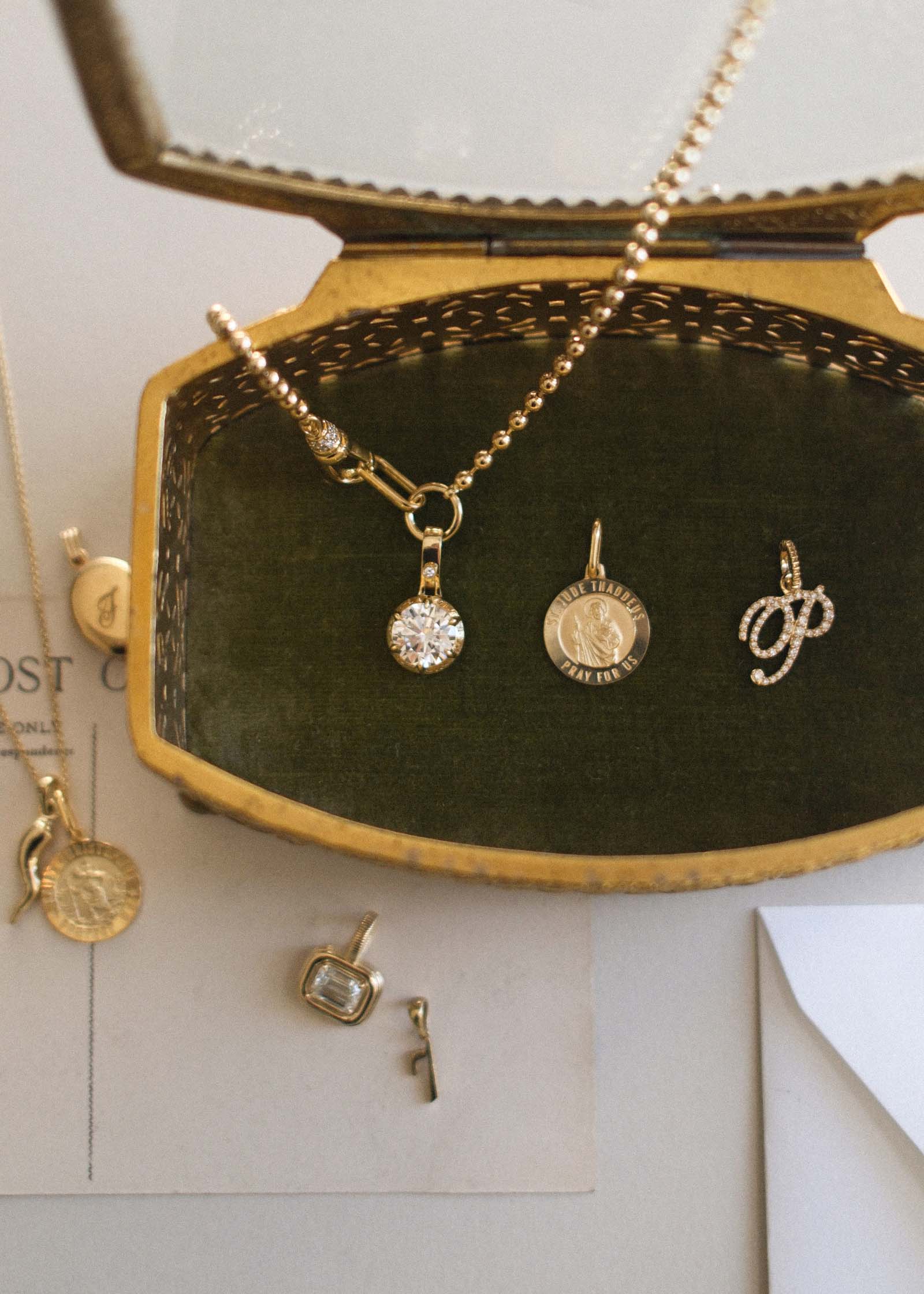 A Kinn 14k Gold Saint Jude Charm Pendant, pave diamond love letter P charm, and a Madeleine Round Diamond Pendant necklace laying in an open gold jewelry box