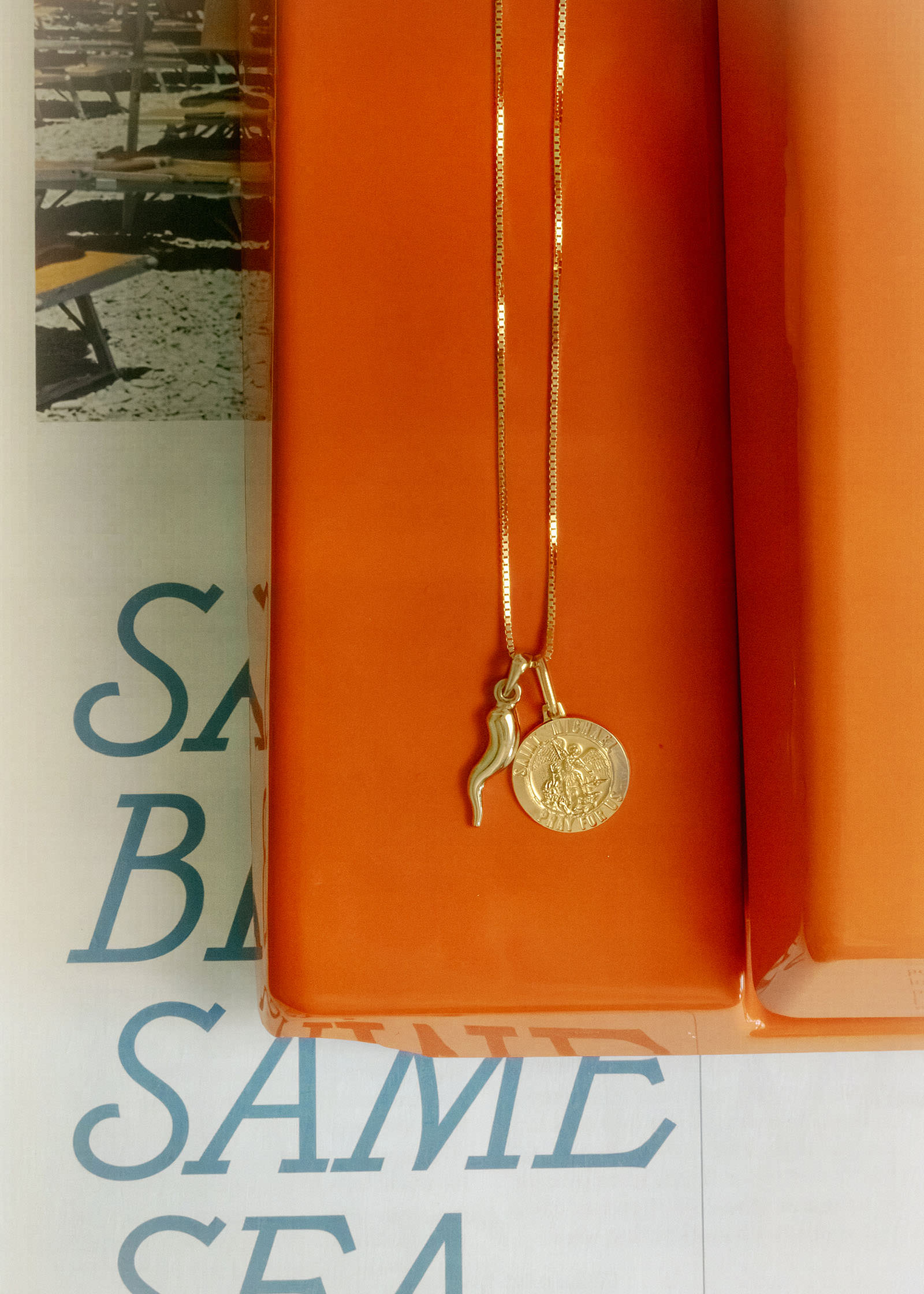 A Kinn 14k Gold Saint Michael Charm Pendant and Alessandra Cornicello Charm  on a Box Chain Necklace laying flat across an orange phone