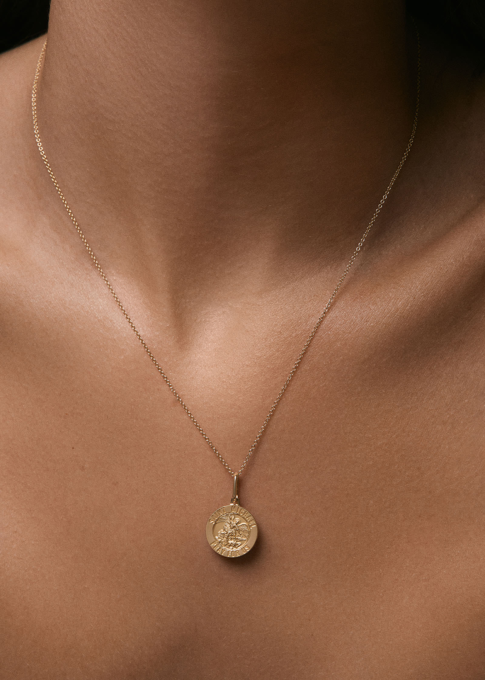 A close up of a woman wearing a Kinn 14k Gold Saint Michael Charm Pendant hanging off a Micro Rolo Chain
