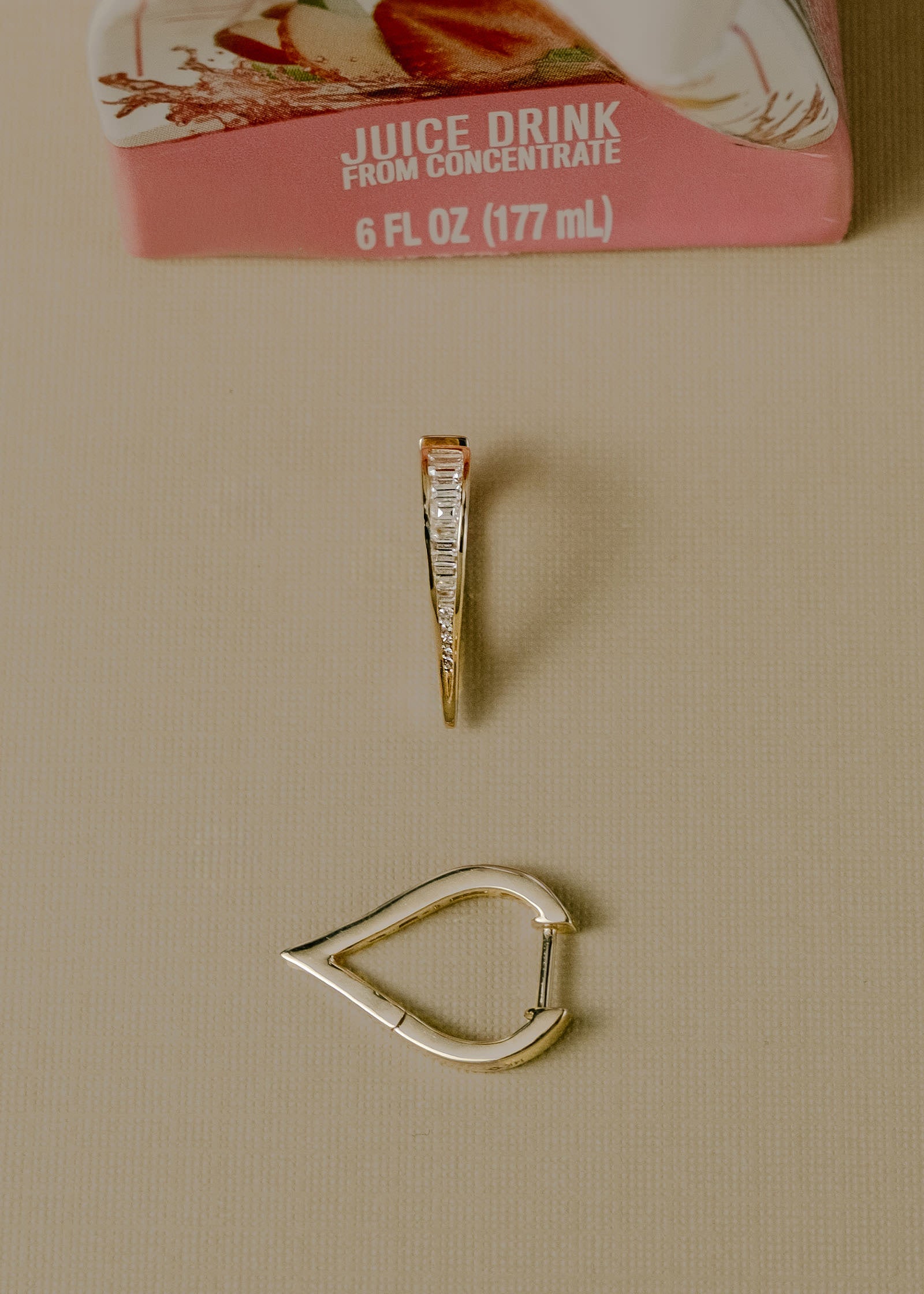 A pair of 14k Gold Viola Baguette Diamond Drop Earrings with one laying facing up and one on its side next to an empty juice box