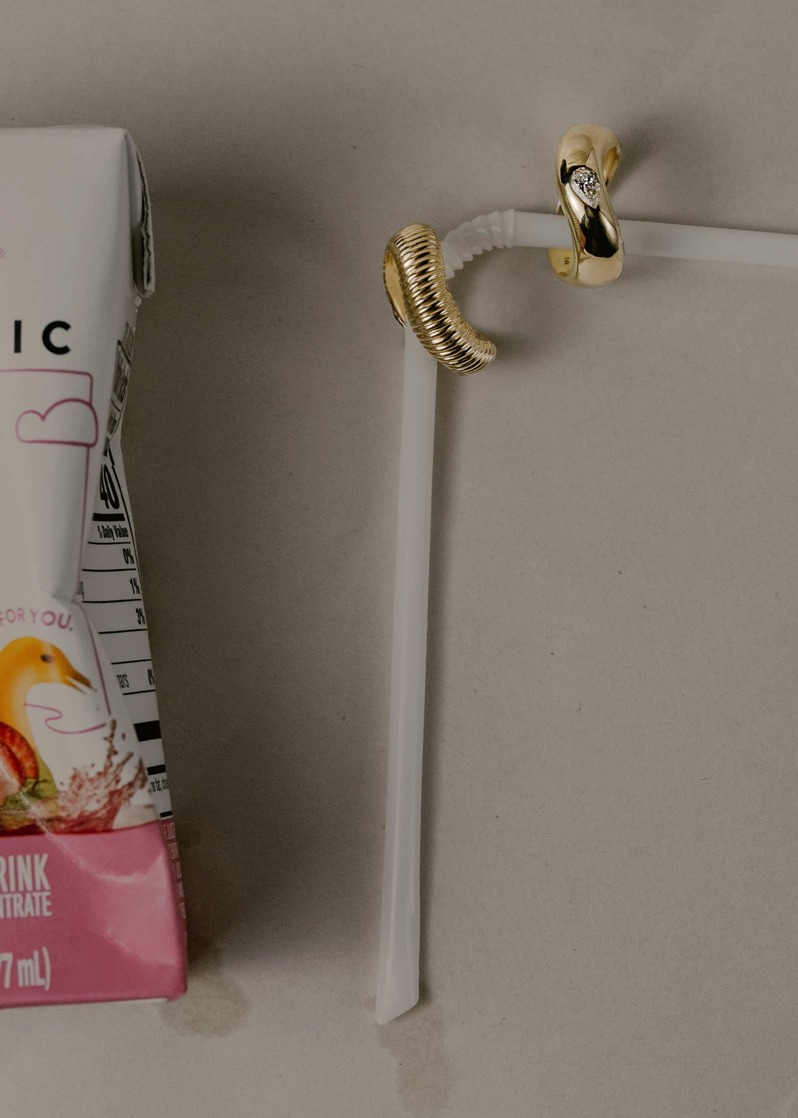 A Kinn 14k Gold Demi Curve Band Ring with a Pear Diamond with a Demi Coil Ribbed Band Ring strung on a straw next to an empty juice box