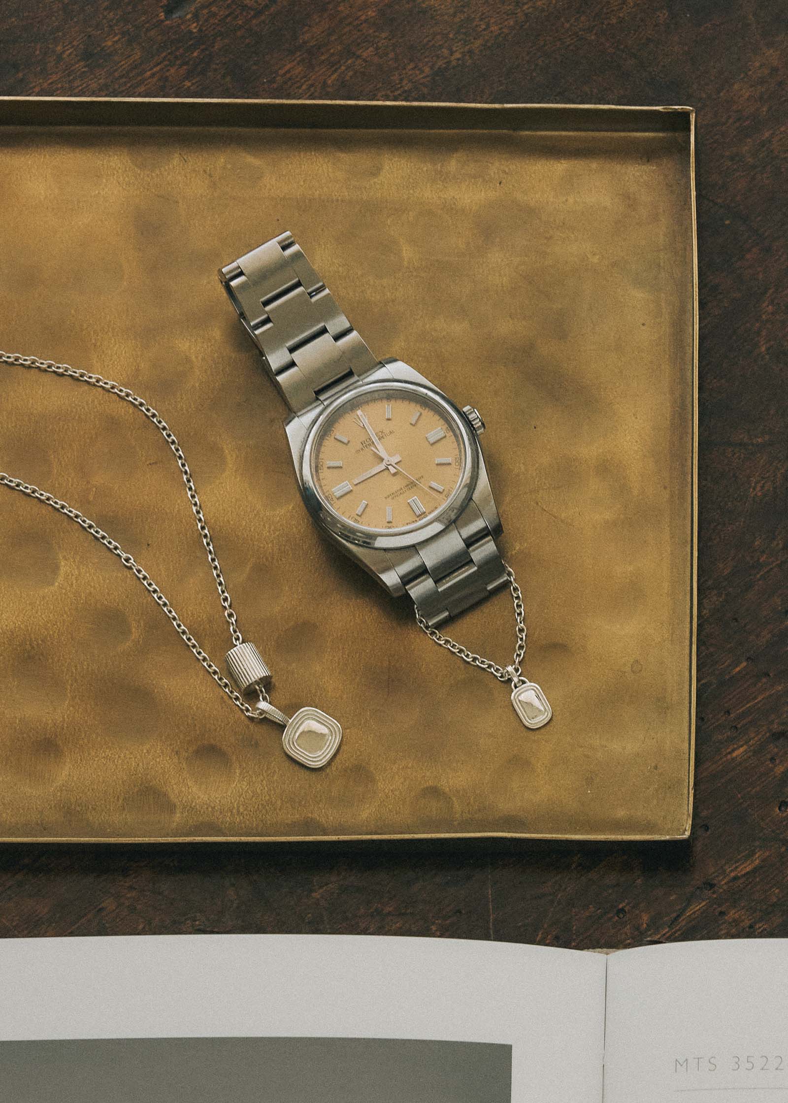 A Kinn x Shoppe Amber Interiors The Estate Pendant and Estate Charm Necklace laying flat on a metal tray next to a metal watch with the Estate Watch Charm attached on the band