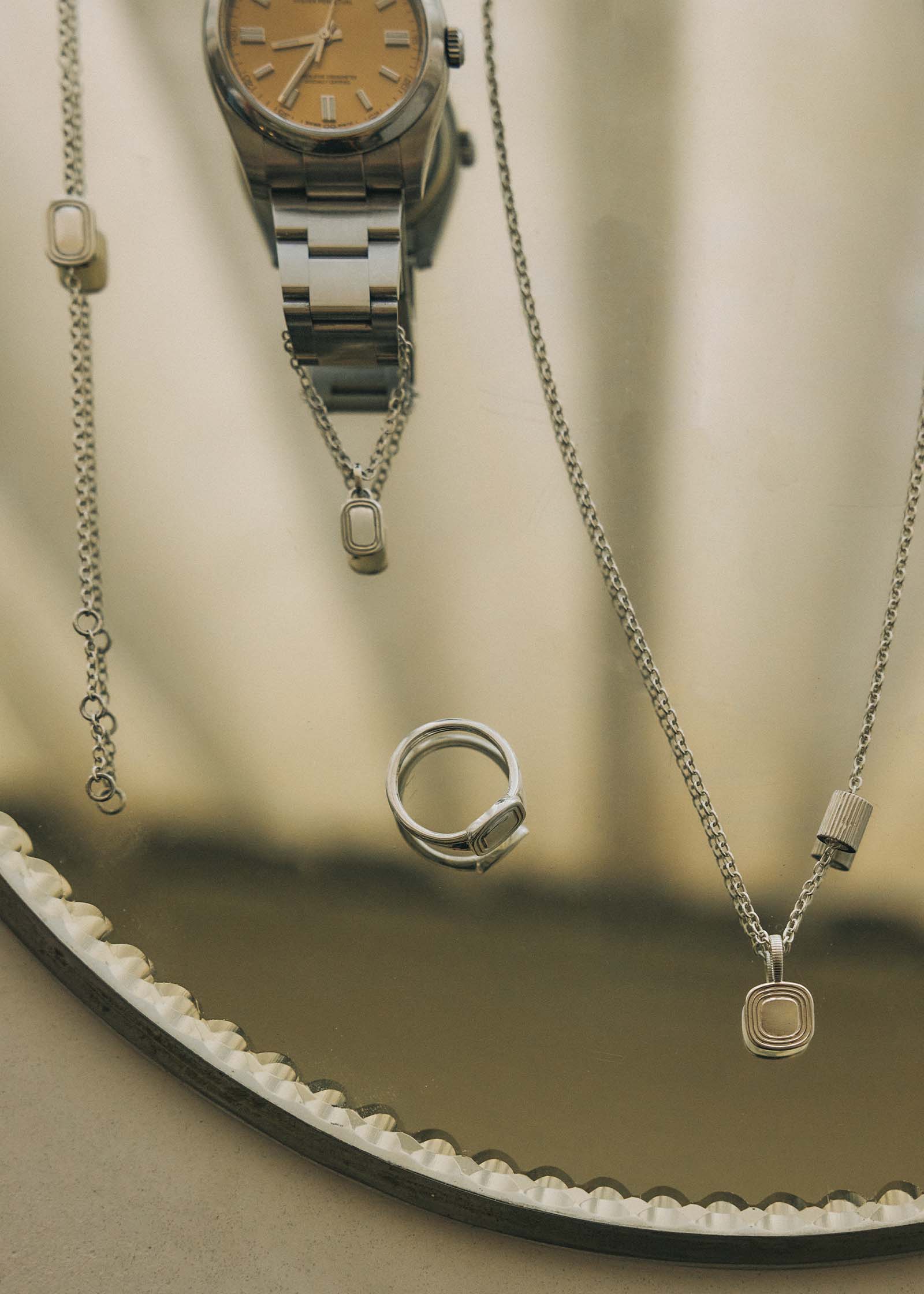 A Kinn x Shoppe Amber Interiors The Estate Pendant and Estate Charm Necklace laying on a mirror next to a metal watch with the Estate Watch Charm attached on the band, The Estate Signet Ring, and The Estate Bracelet all in 925 sterling silver