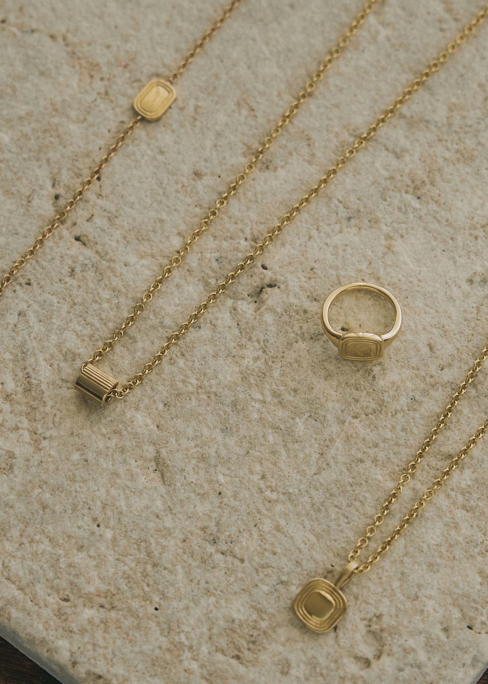  A Kinn x Shoppe Amber Interiors The Estate Charm 14k Yellow Gold on a cable rolo chain laying flat with The Estate Bracelet, Estate Signet Ring, and the Estate Necklace on a stone slab