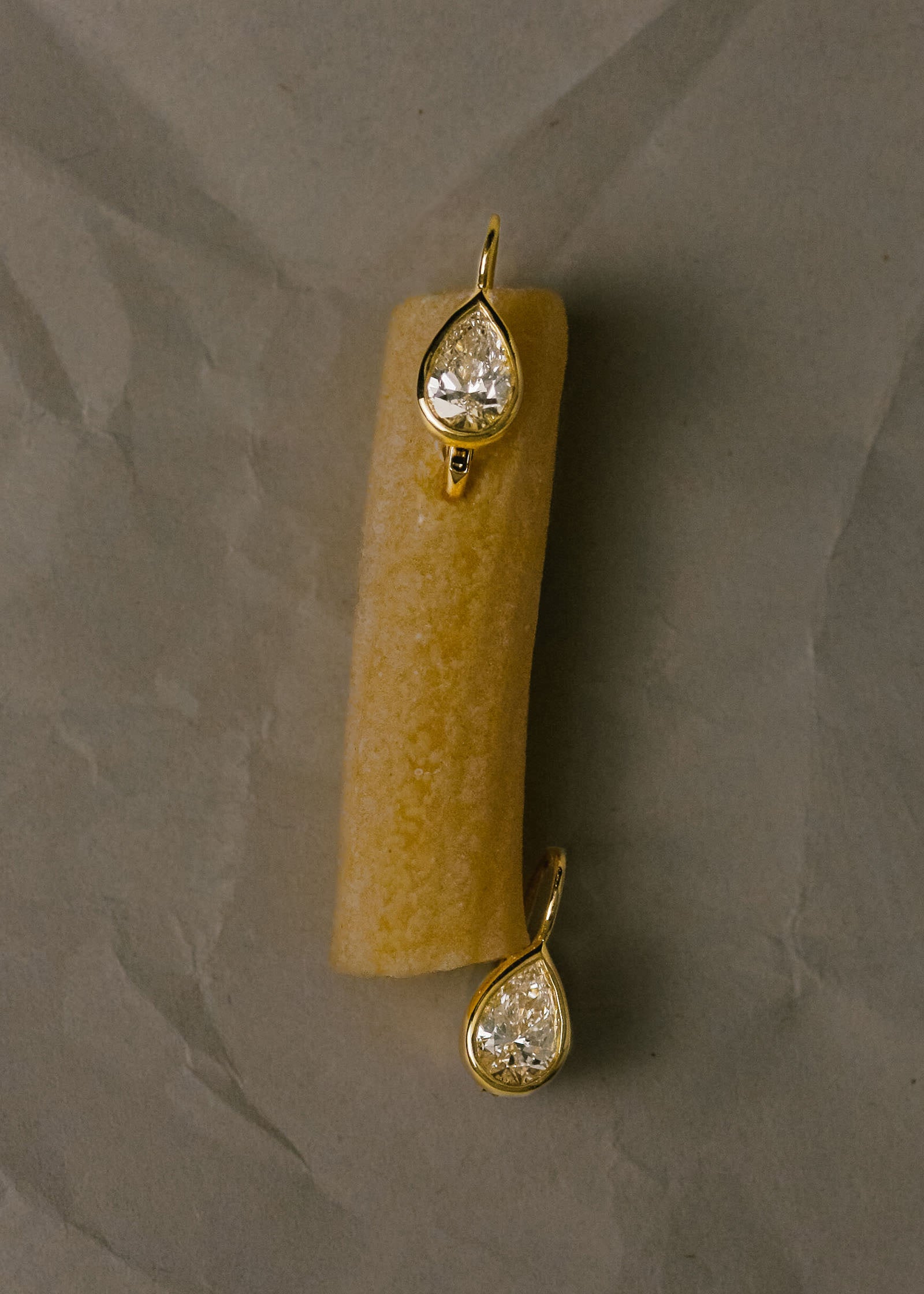 A pair of Kinn 14k Gold 2ctw Pear Diamond Drop Earrings laying on a piece of penne pasta against a crinkled dark gray background