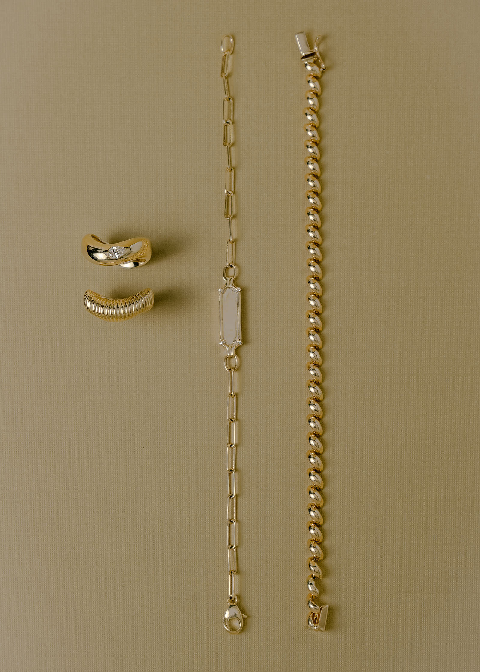a Kinn 14k Yellow Gold Petite Hampshire House Bracelet laying flat with a Harlow Pave ID Bracelet and a Demi Curve & Coil Ring Stack against a beige background