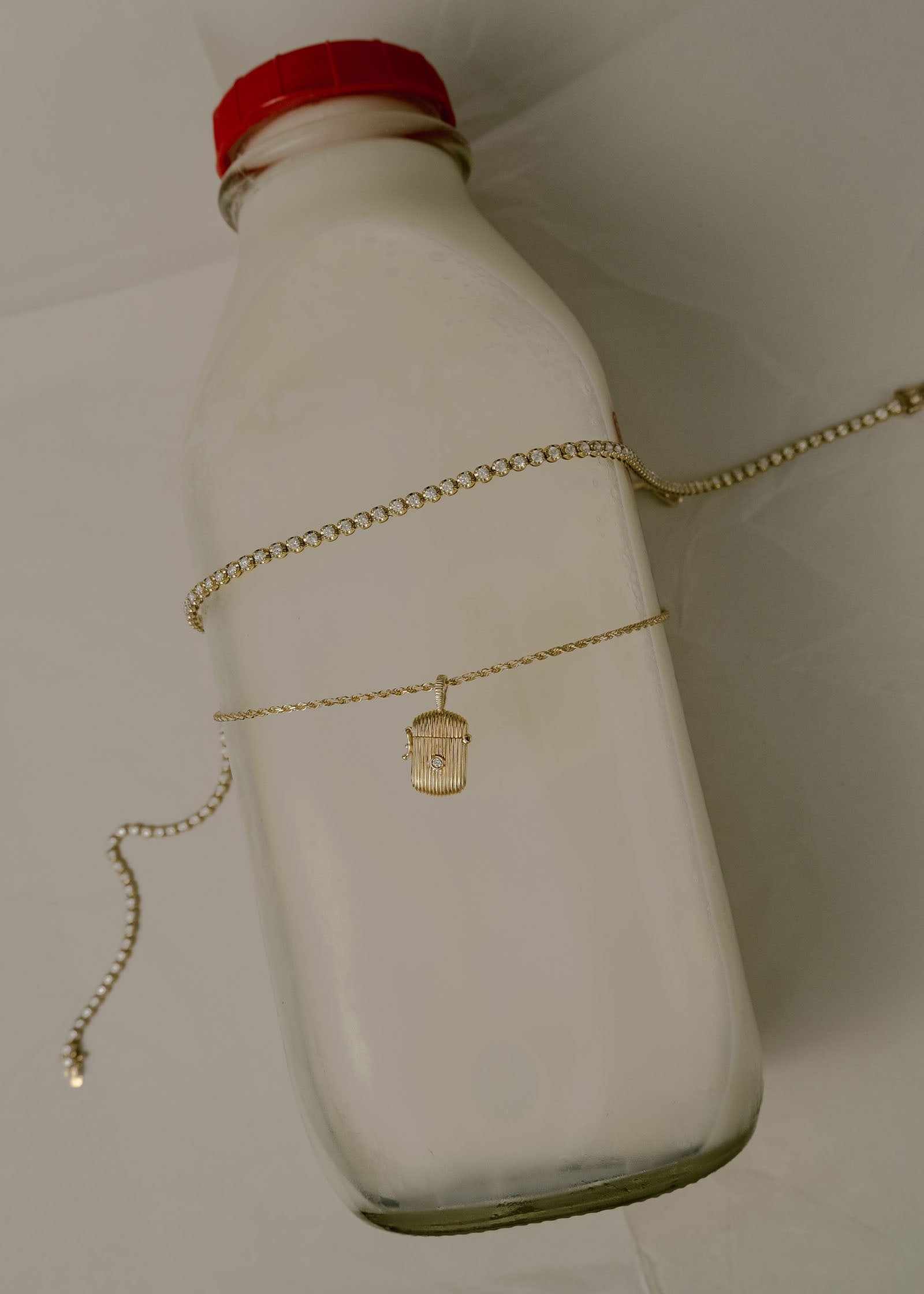 a Kinn 14k Yellow Gold Madison Diamond Tennis Necklace laying across a bottle of milk with a Relic Ribbed Locket Necklace