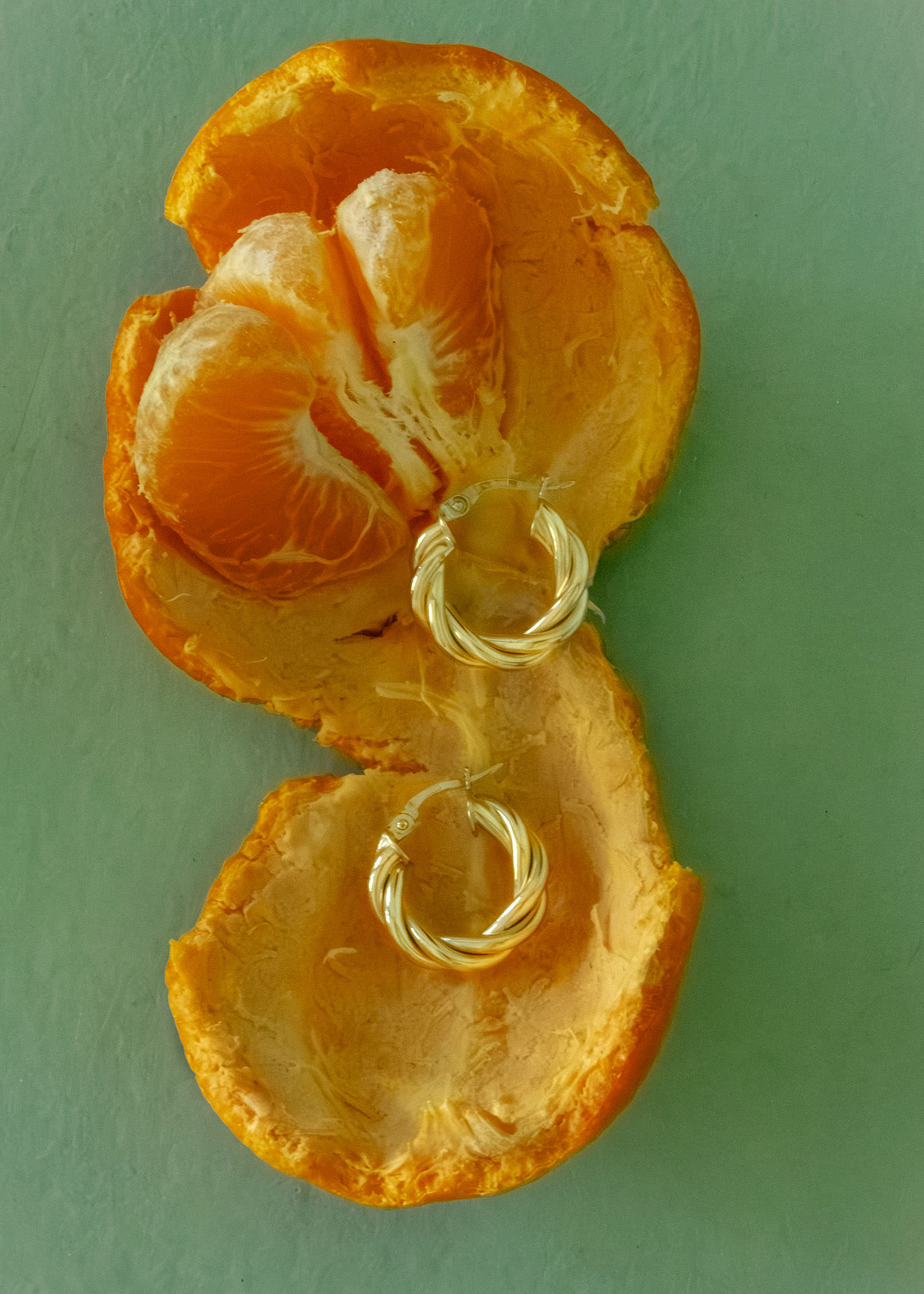 A pair of Kinn 14k Gold Taylor Twist Petite Hoop Earrings laying flat a peeled open orange against a green background