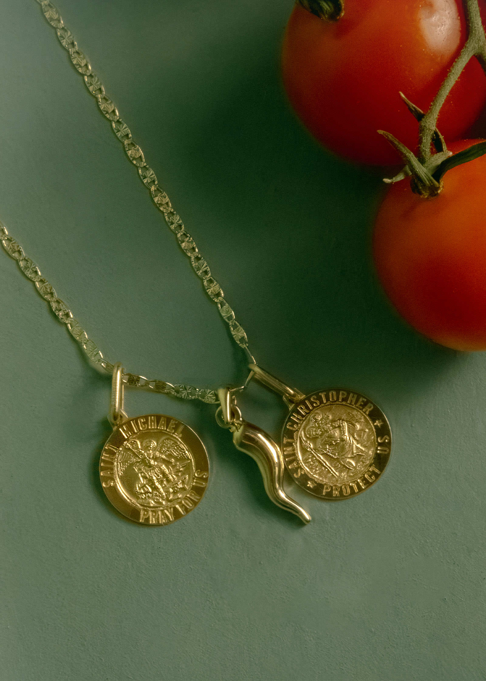 A Kinn 14k Valentino Tri-Color Gold Chain Necklace with a Saint Michael Pendant, Saint Christopher Pendant, and a Alessandra Cornicello charm laying flat against a green background next to a couple tomatoes