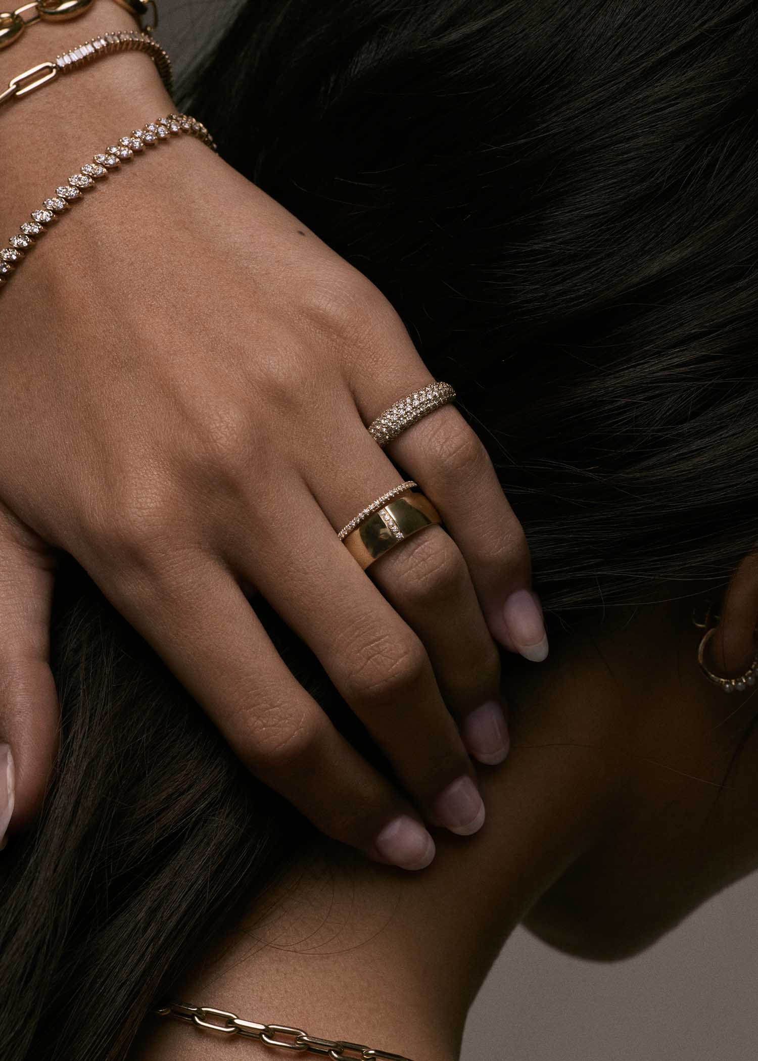 woman's hand resting against her head wearing a Kinn 14k Gold 1.2mm Dahlia Pavé Diamond Band Ring and Vertical Pave Cigar Band stacked on her ring finger and a Scarlett Pavé Diamond Dome Ring on her pink finger