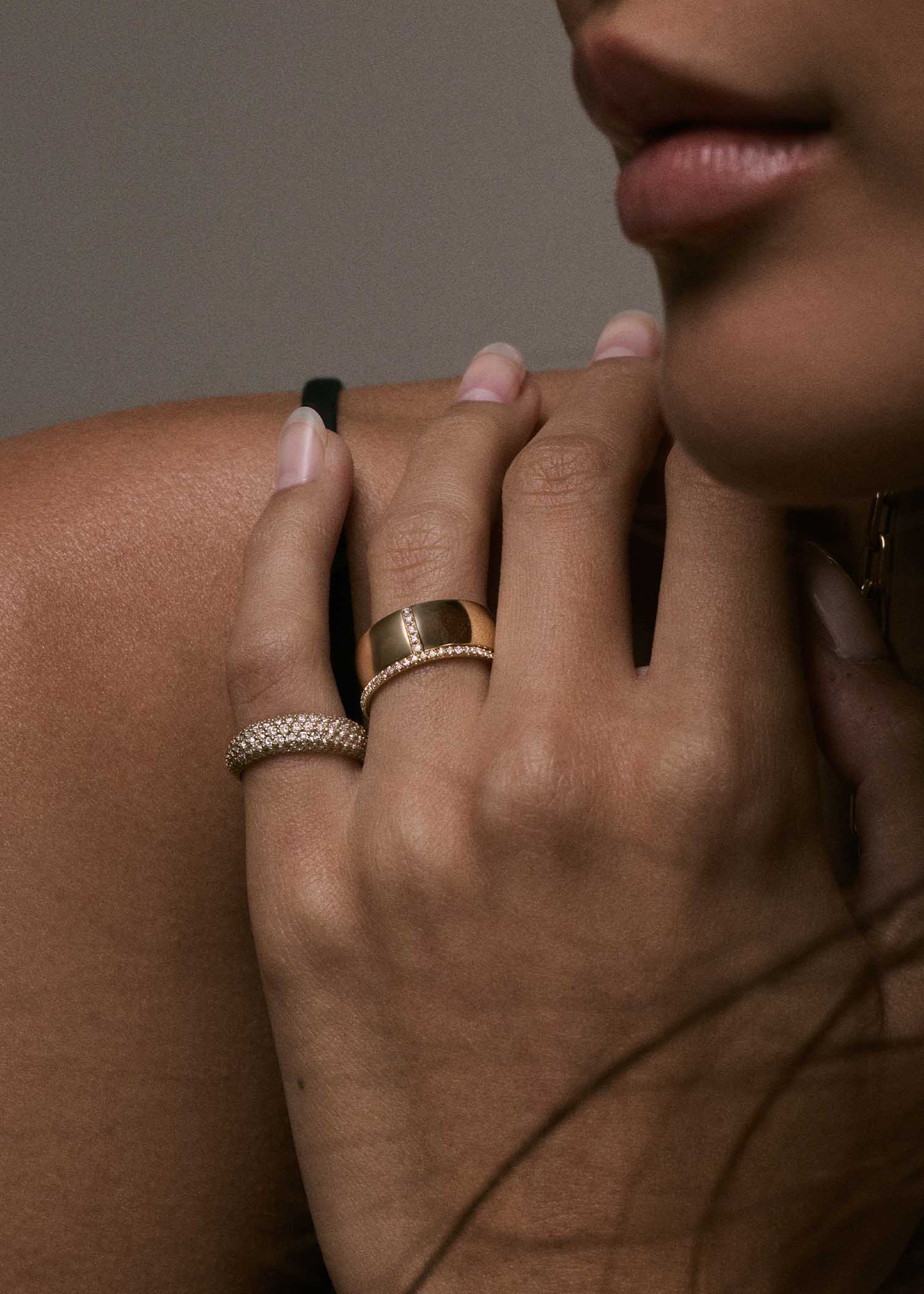 woman's hand resting against her shoulder wearing a Kinn 14k Gold 1.2mm Dahlia Pavé Diamond Band Ring and Vertical Pave Cigar Band stacked on her ring finger and a Scarlett Pavé Diamond Dome Ring on her pink finger