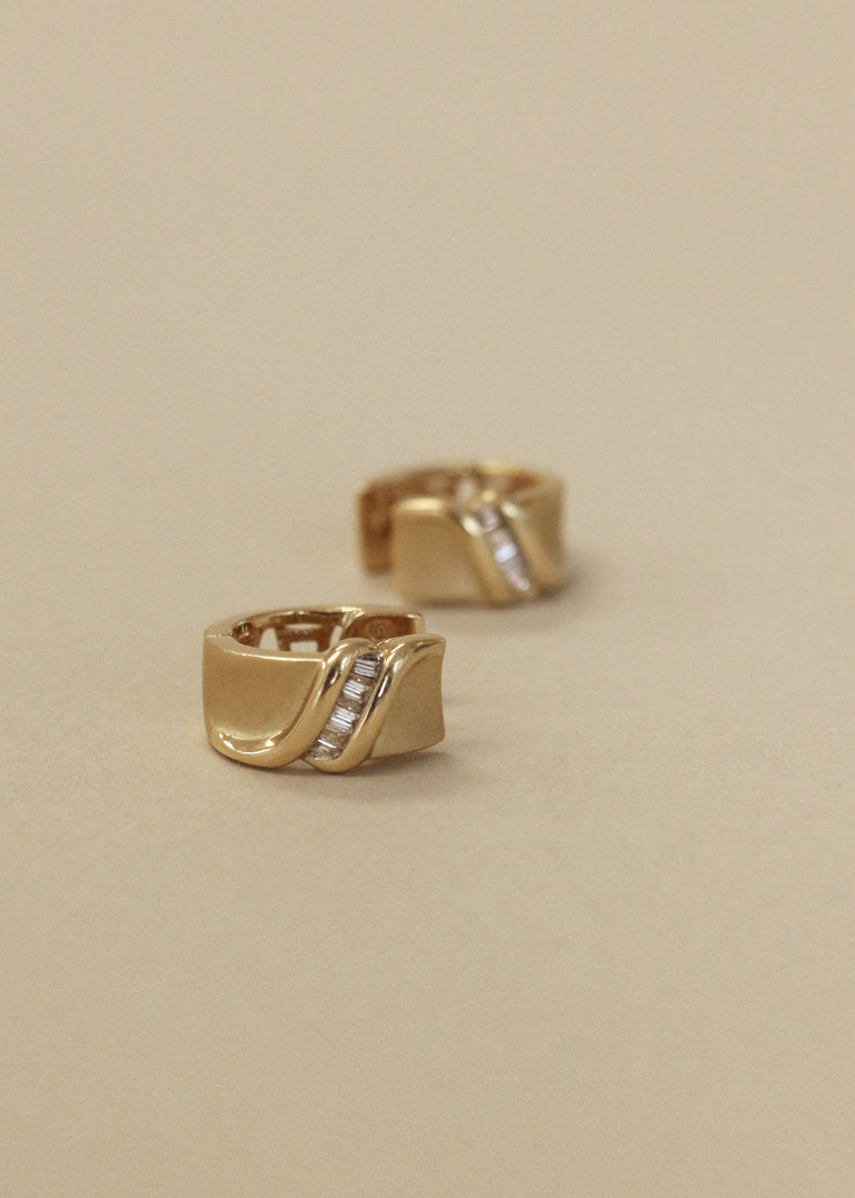 A pair of Kinn Vintage 14k Gold Baguette Diamond Huggie Earrings laid flat on its side